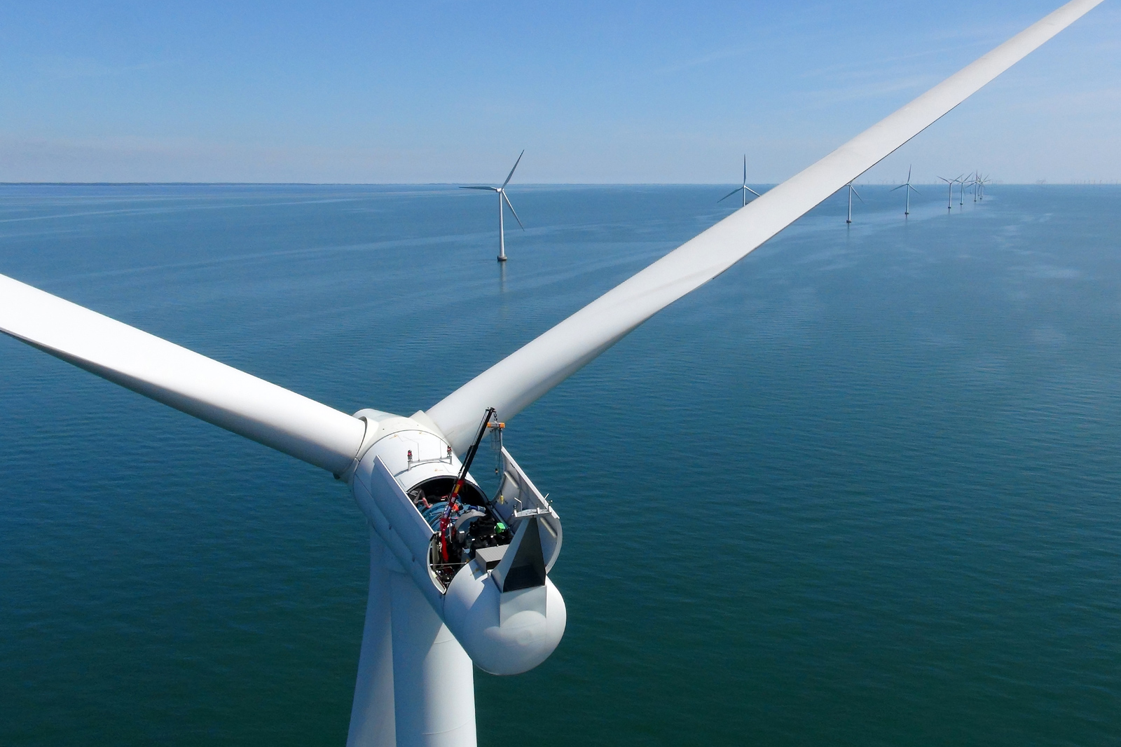 A close-up view of an offshore wind turbine with a calm sea and additional turbines visible in the background under a clear blue sky.