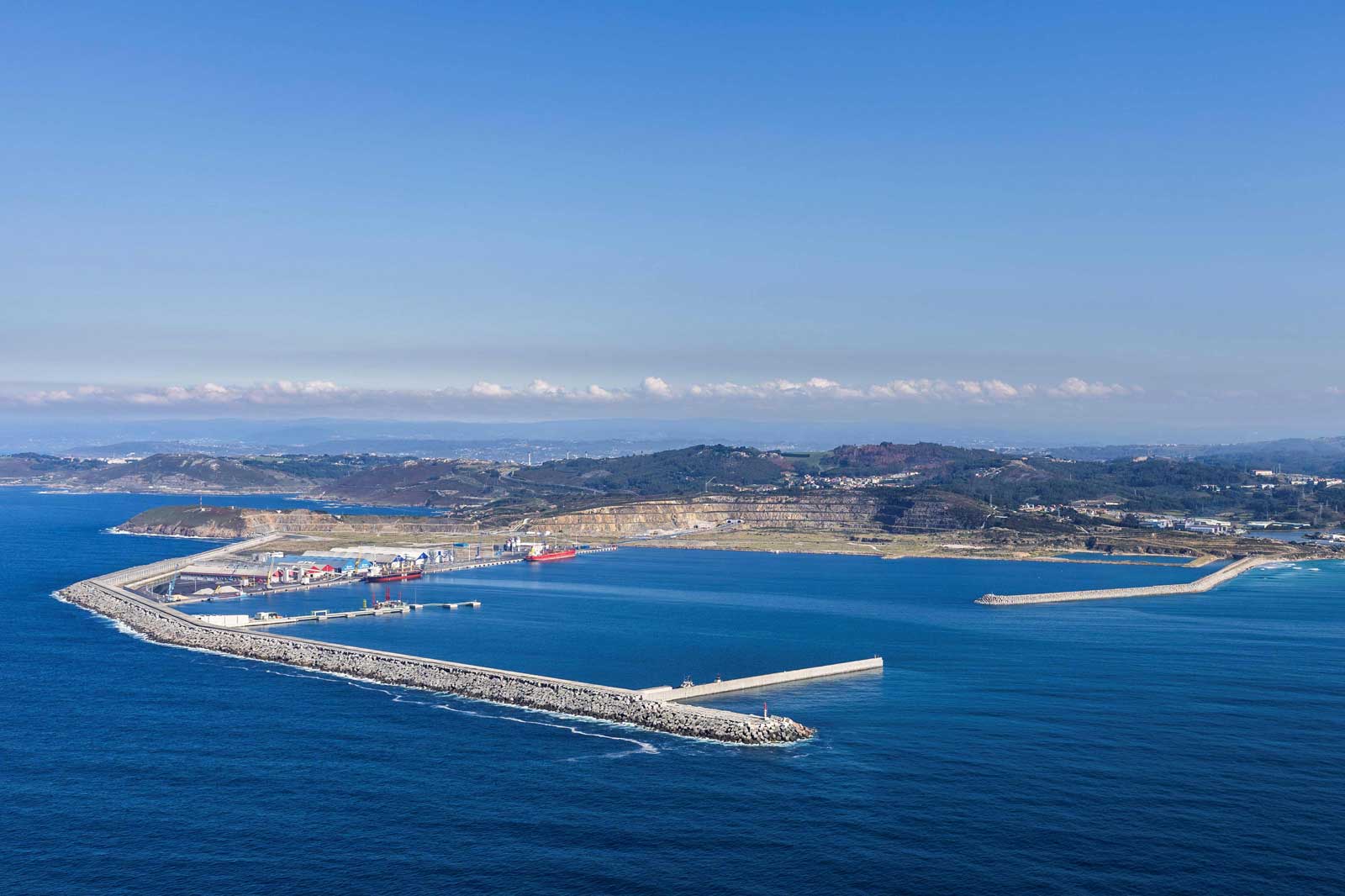 Aerial view of a coastal landscape with a long pier, ships at port, and clear blue water under a bright sky.