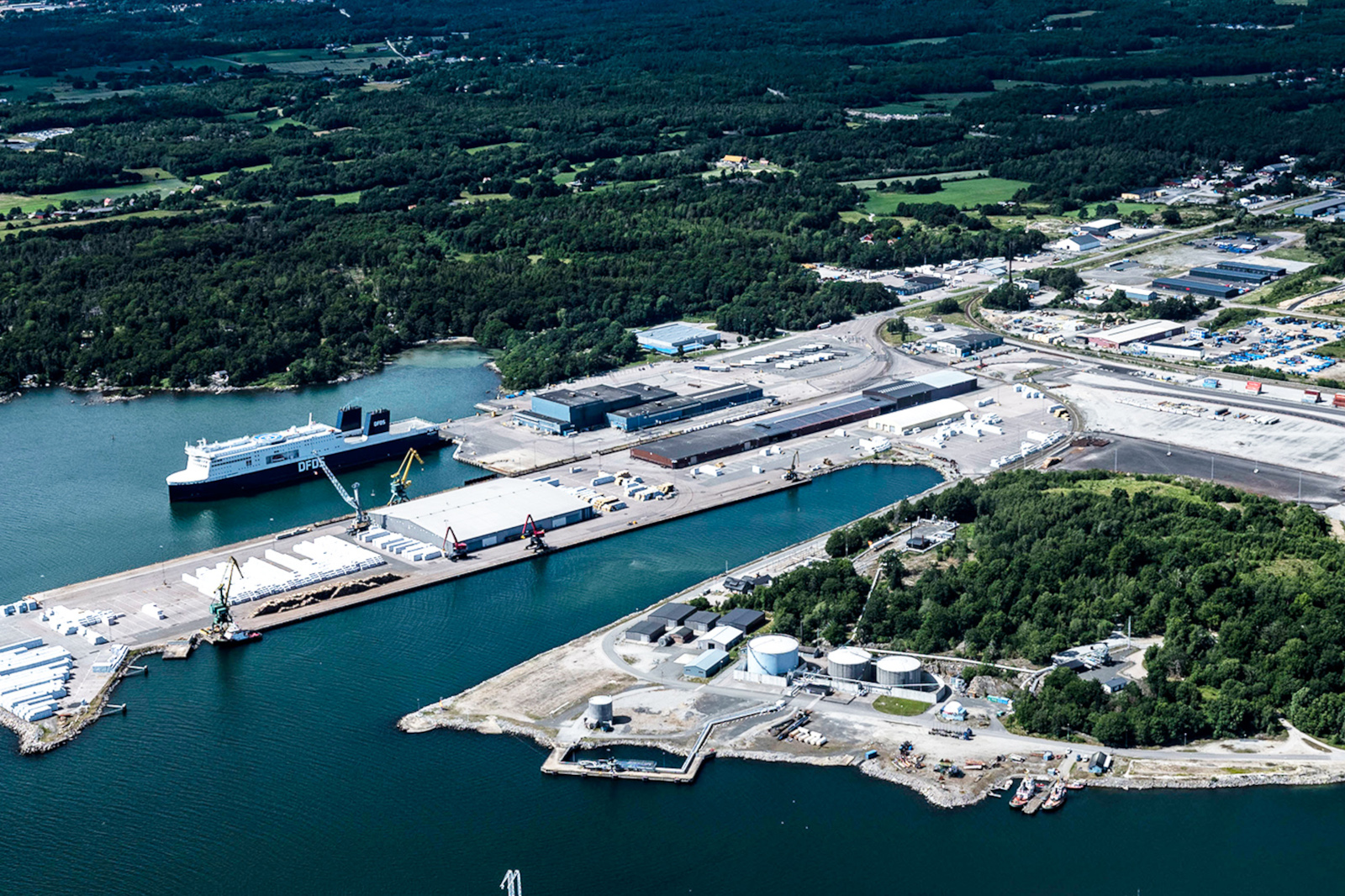 Aerial view of a busy port with a ferry docked, surrounded by green trees and industrial buildings.