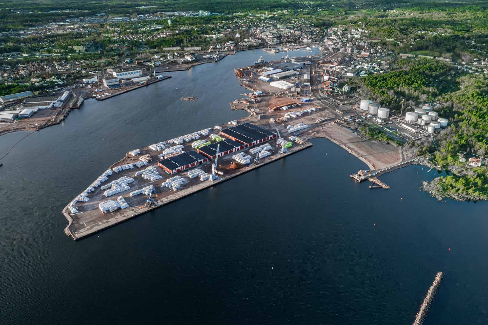 An aerial view of a harbour with warehouses, boats, and industrial facilities along the waterfront, surrounded by greenery.