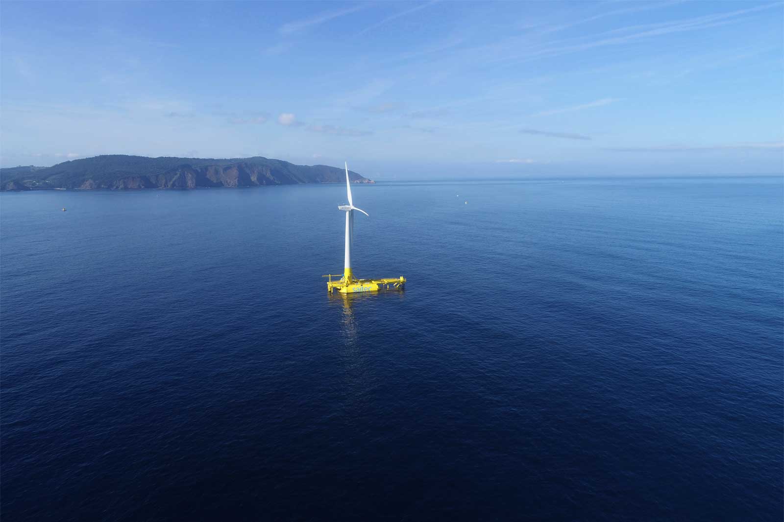 A floating wind turbine in calm blue waters with a mountainous coastline in the background under a clear sky.