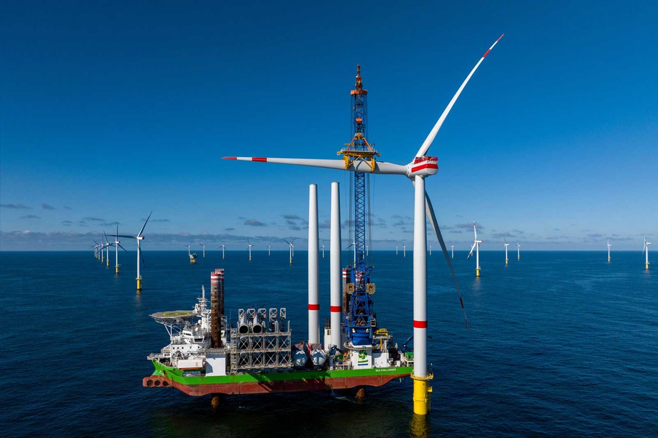 Ein großes Offshore-Windkraftanlageninstallationsschiff mit mehreren Windkraftanlagen im Hintergrund unter einem klaren blauen Himmel.