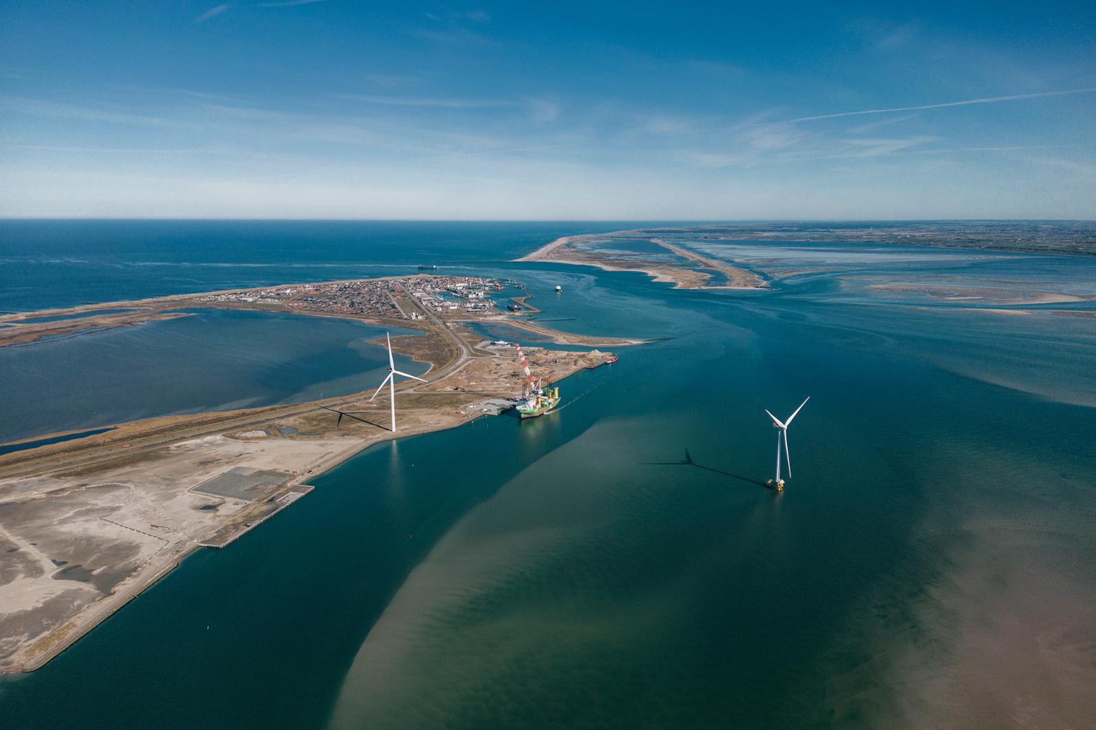 Aerial view of wind turbines in a coastal area, with a town, sandy shores, and calm waters under a clear blue sky.