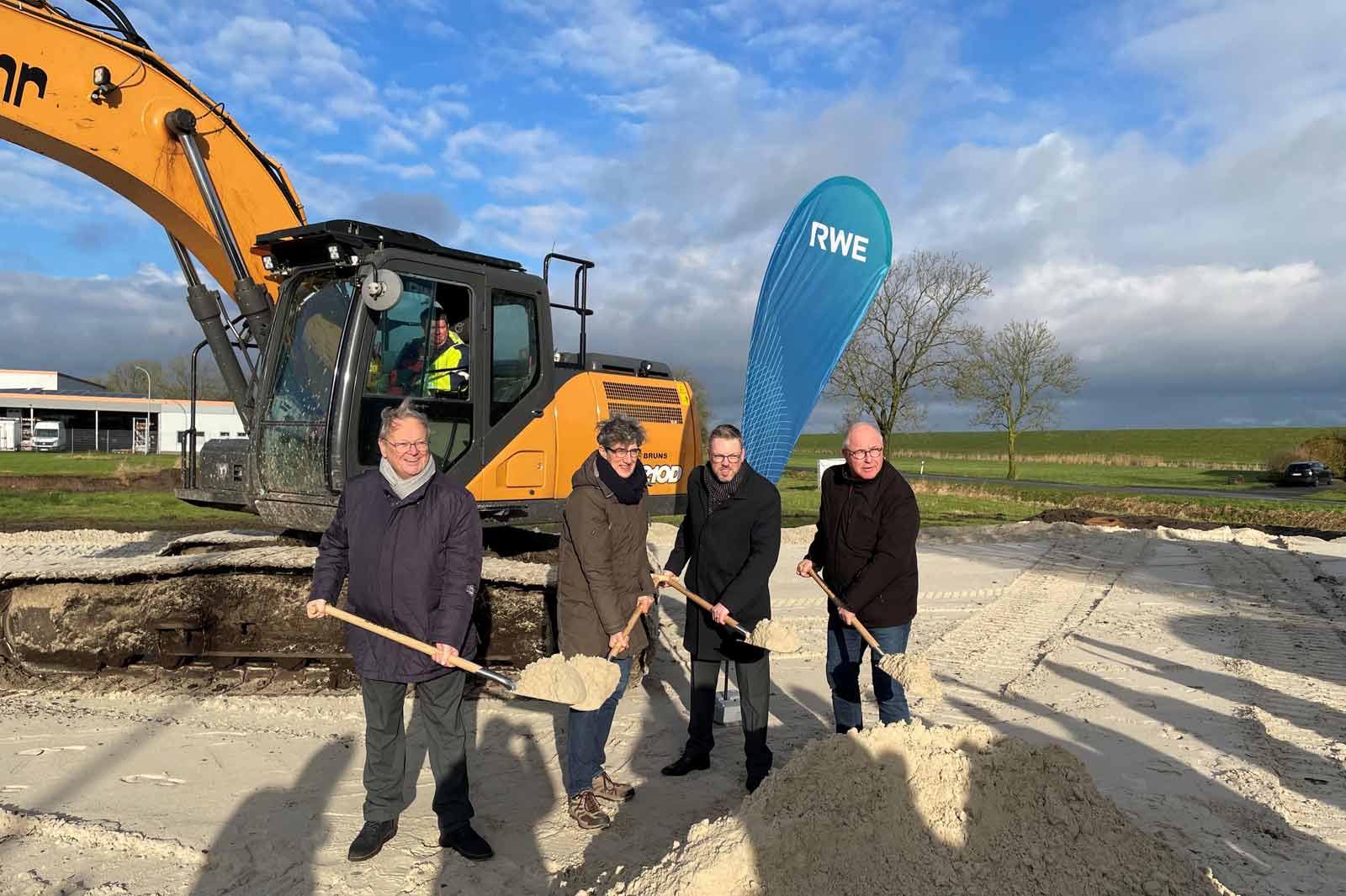 Menschen nehmen an einer Grundsteinlegung mit einem Bagger und einer blauen RWE-Flagge in einer ländlichen Landschaft teil.