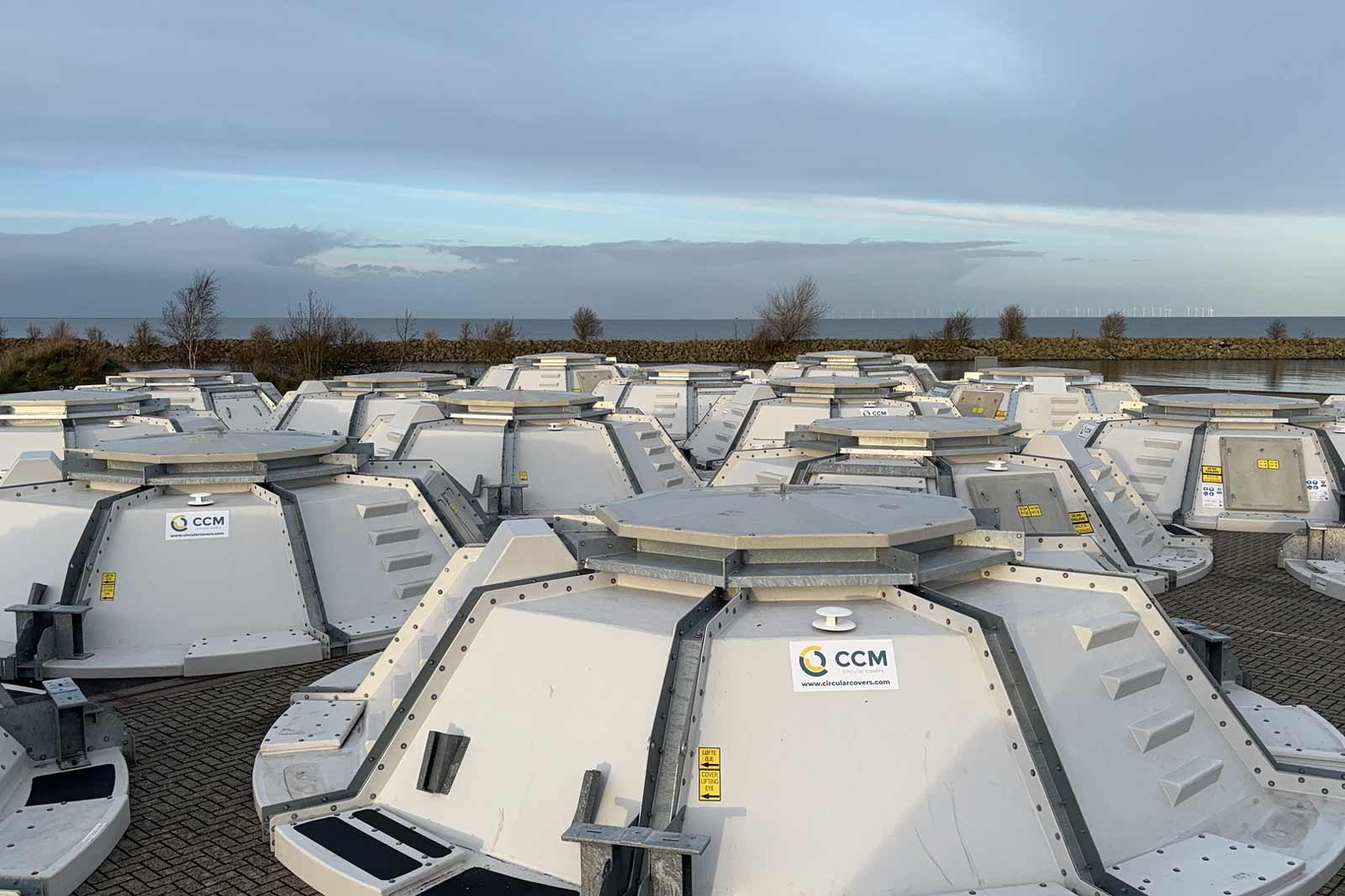 Several circular covers in white and grey are arranged on a paved area, with a view of the sea in the background.
