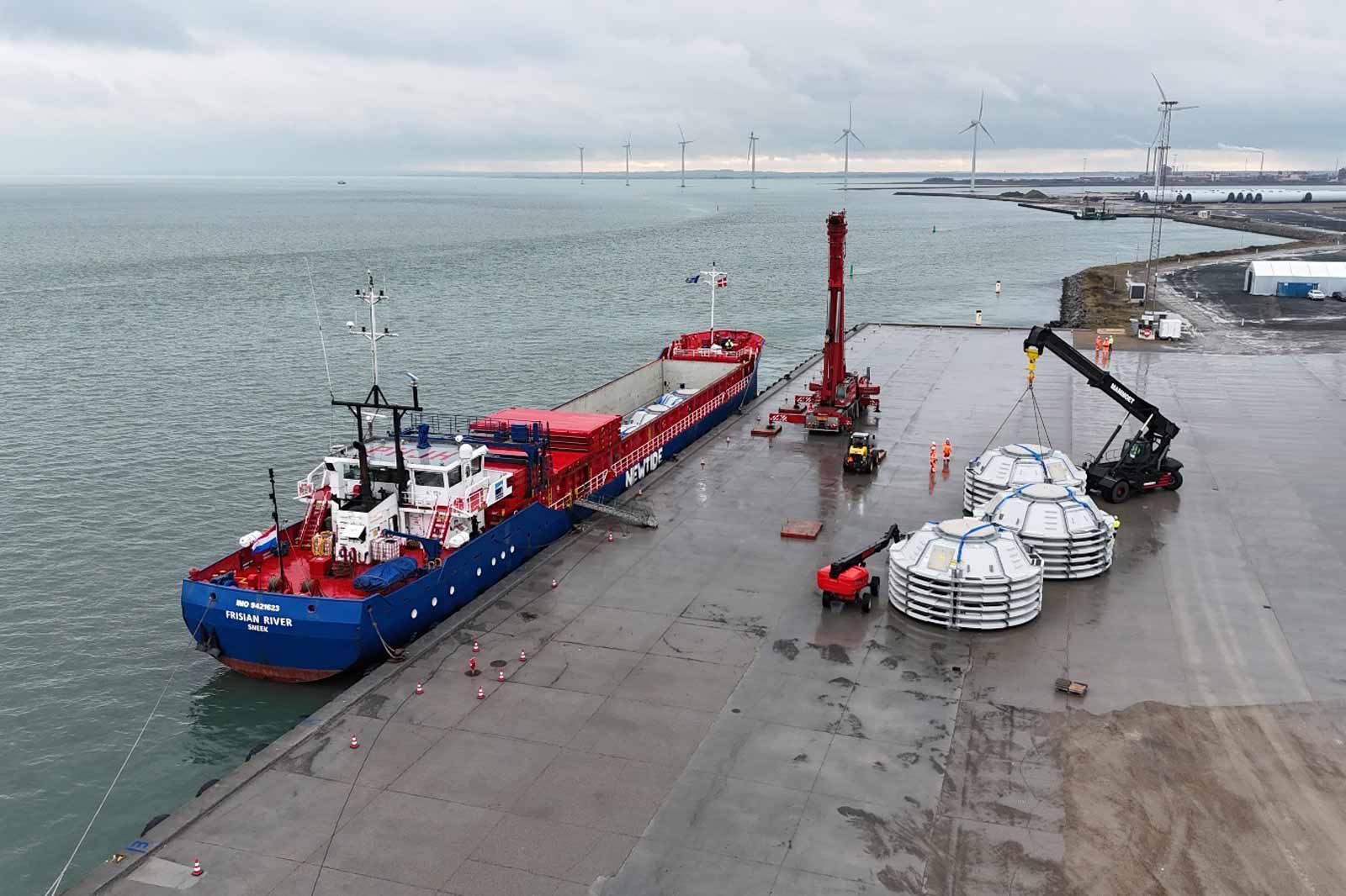 A cargo ship named Frisian River docked at the quay, with wind turbines in the background and workers loading large materials.