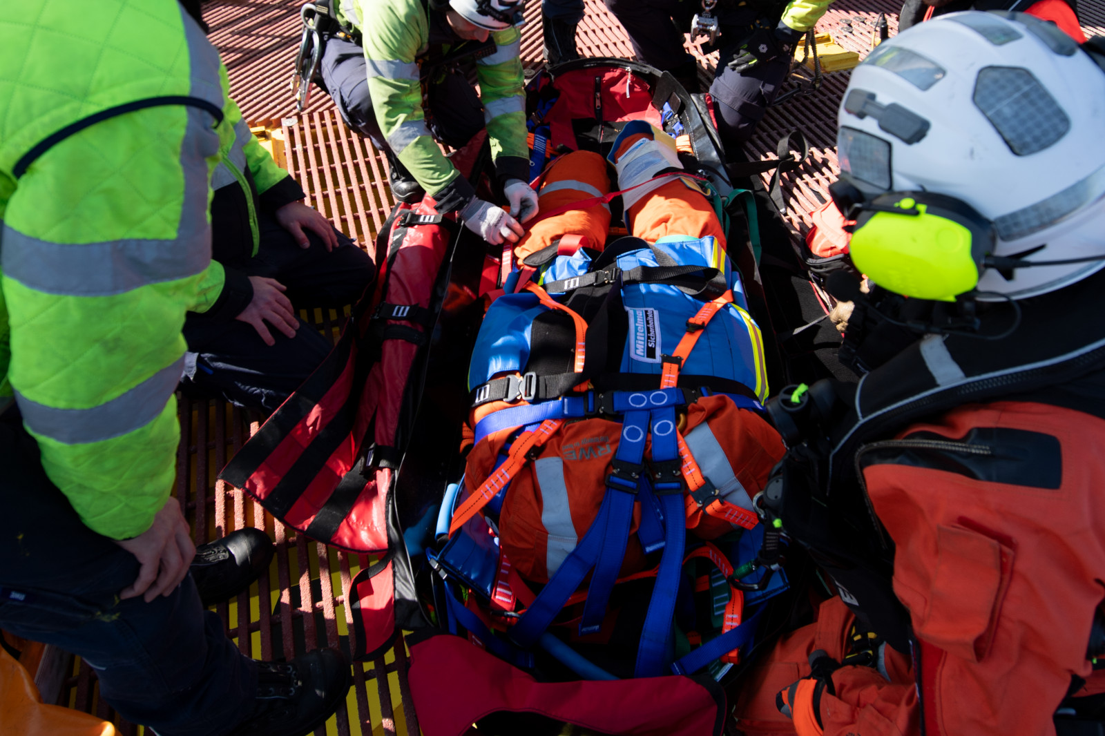 Rettungskräfte arbeiten zusammen, um eine verletzte Person in einem orange-blauen Sicherheitsanzug auf einer Trage zu stabilisieren.
