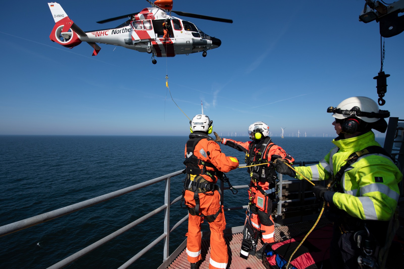 Ein Rettungshubschrauber schwebt über dem Wasser, während zwei Einsatzkräfte mit orangenen Anzügen sich auf Rettungsaktionen vorbereiten.