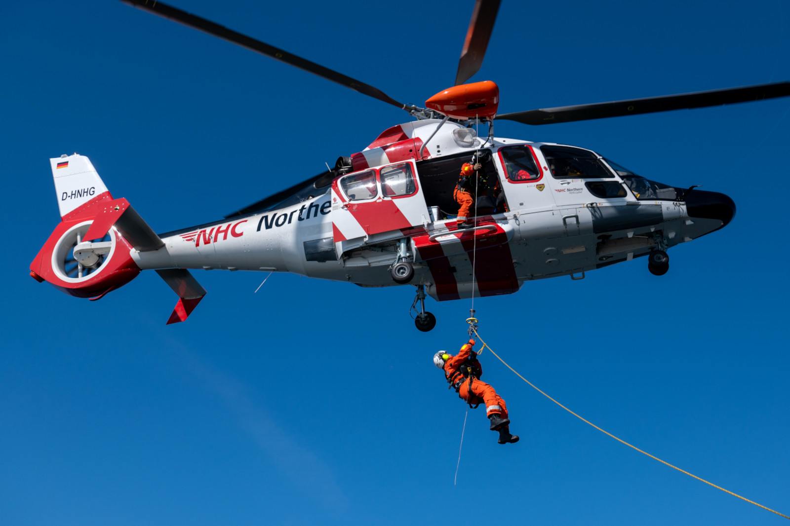 Ein Hubschrauber mit der Aufschrift 'NHC Northern' schwebt über blauen Himmel, während ein Retter an einem Seil abgelassen wird.