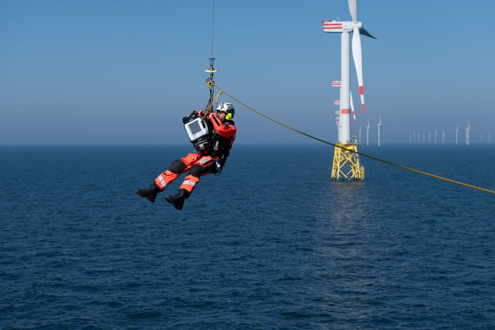 Ein Rettungssanitäter schwebt über dem Meer, während er an einem Seil von einem Windkraftwerk herabgelassen wird.