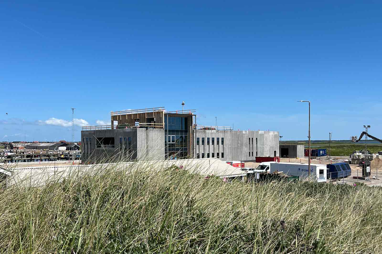 A construction site featuring a modern concrete and glass building by the sea, with grass in the foreground.
