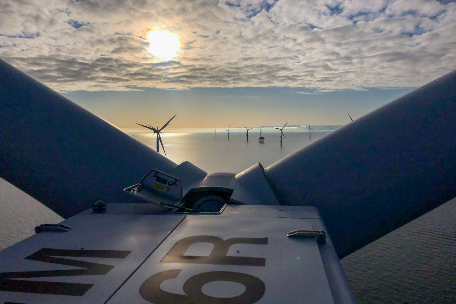 A view from the top of a wind turbine overlooking a sea filled with wind farms under a cloudy sky and bright sun.