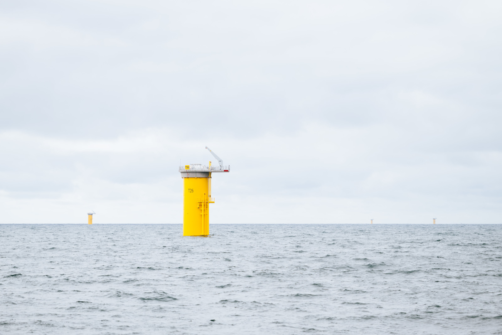 Yellow offshore wind turbine structures rise from the calm sea under a cloudy sky. The nearest one is labelled T26.