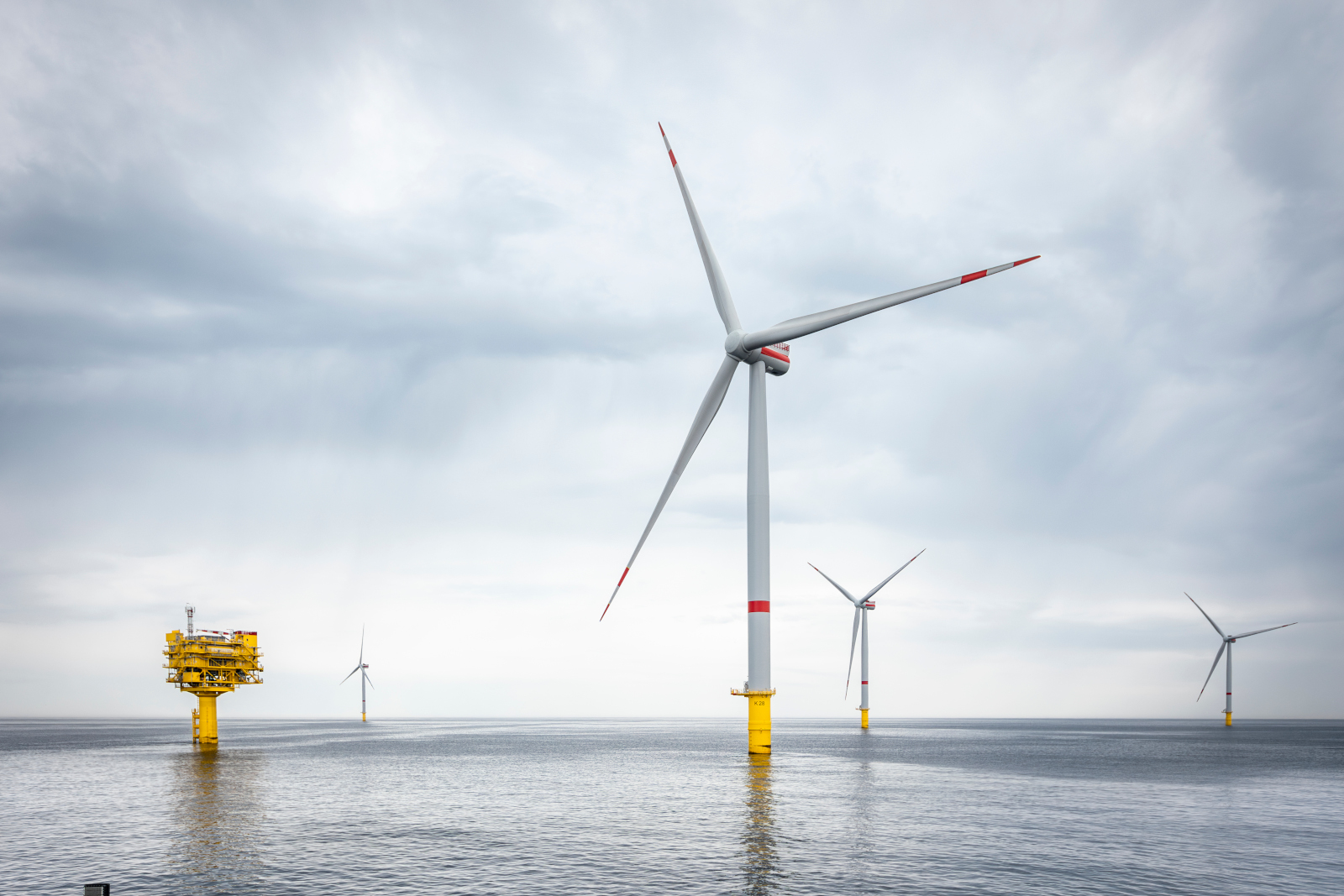 Several wind turbines stand in the water under a grey sky, with a reflective water surface. An offshore platform is visible.