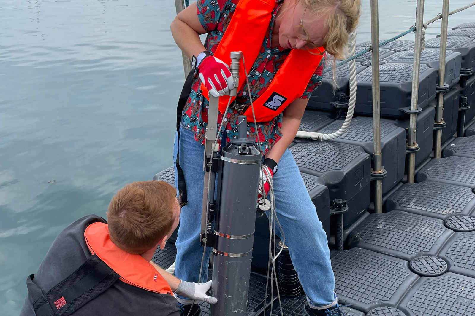 A person in an orange life jacket is holding a device on a boat while another individual observes the action.