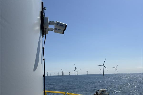 The image shows an offshore wind farm. The view is from a platform over the sea, with wind turbines in the background.