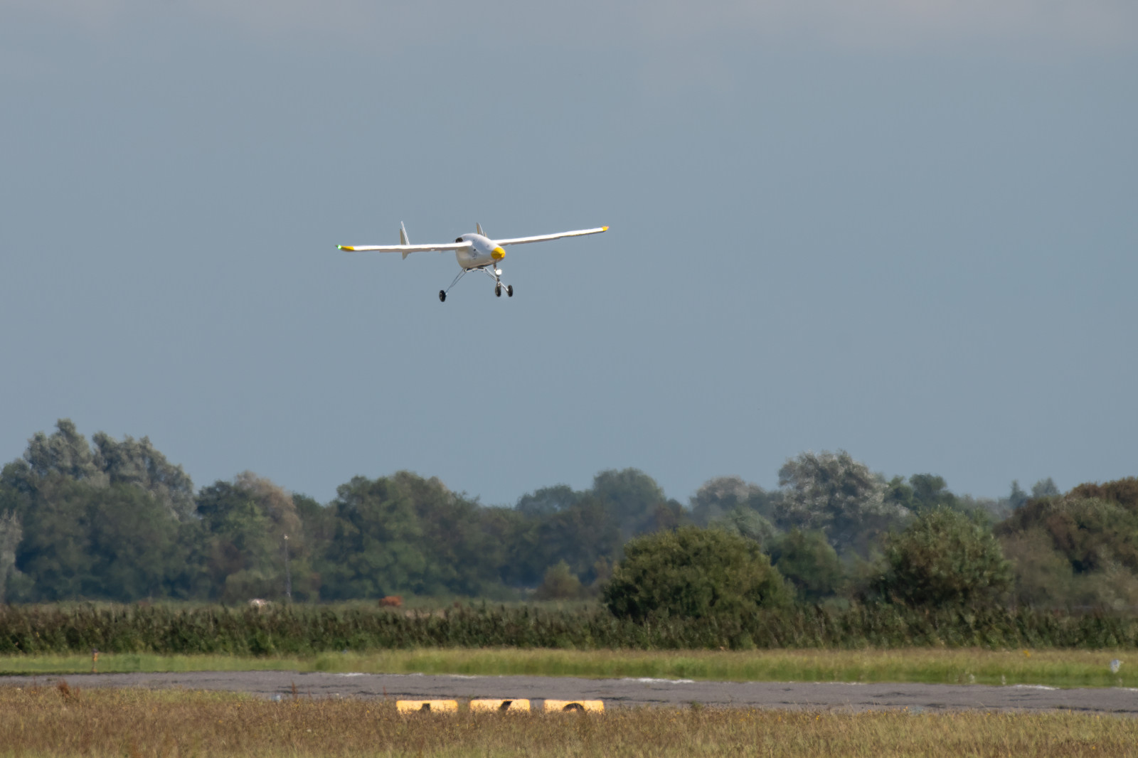 A drone with yellow and green markings is landing over a lush landscape under a clear sky.