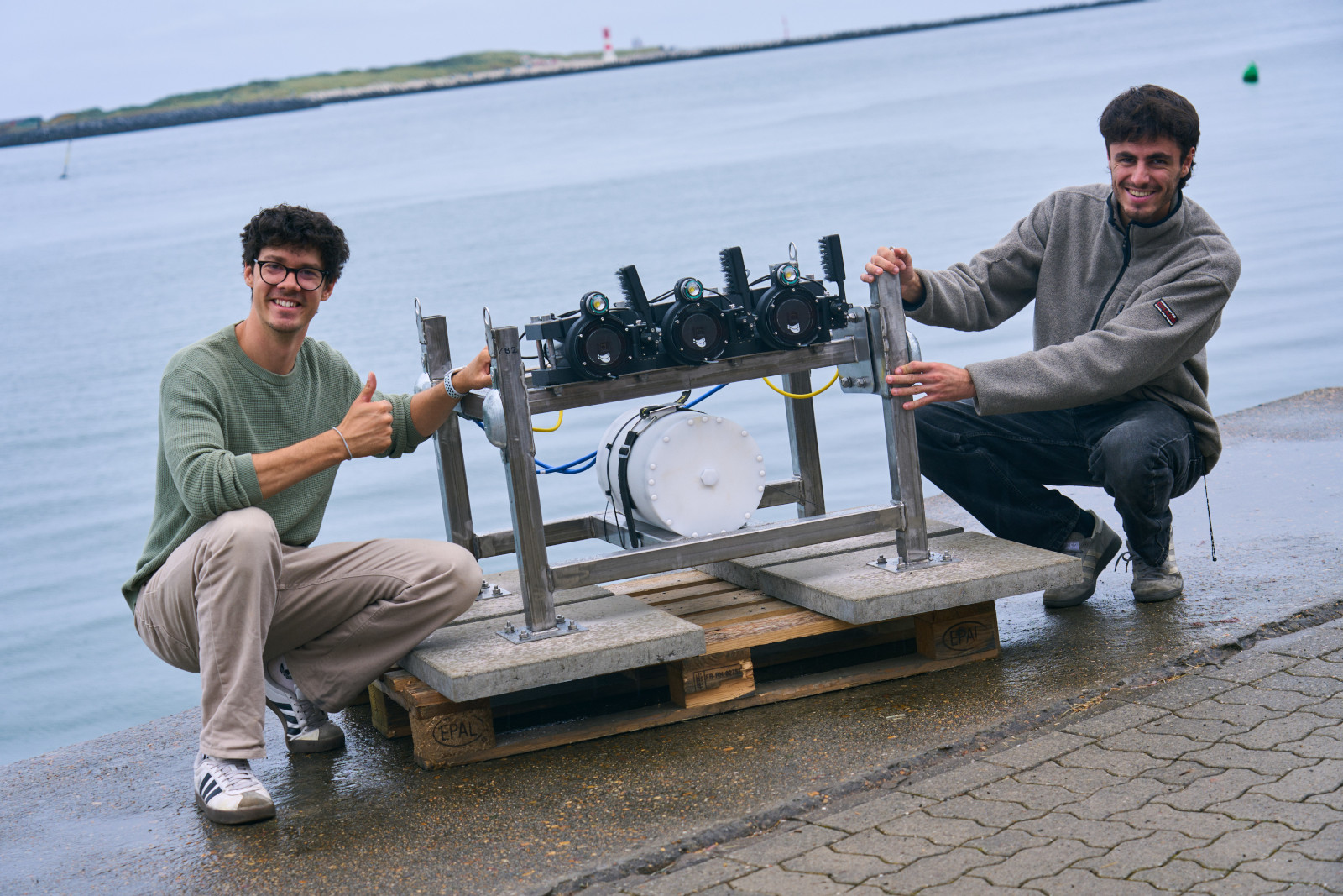 Two individuals showcase a technical device with cameras, placed along a waterway.