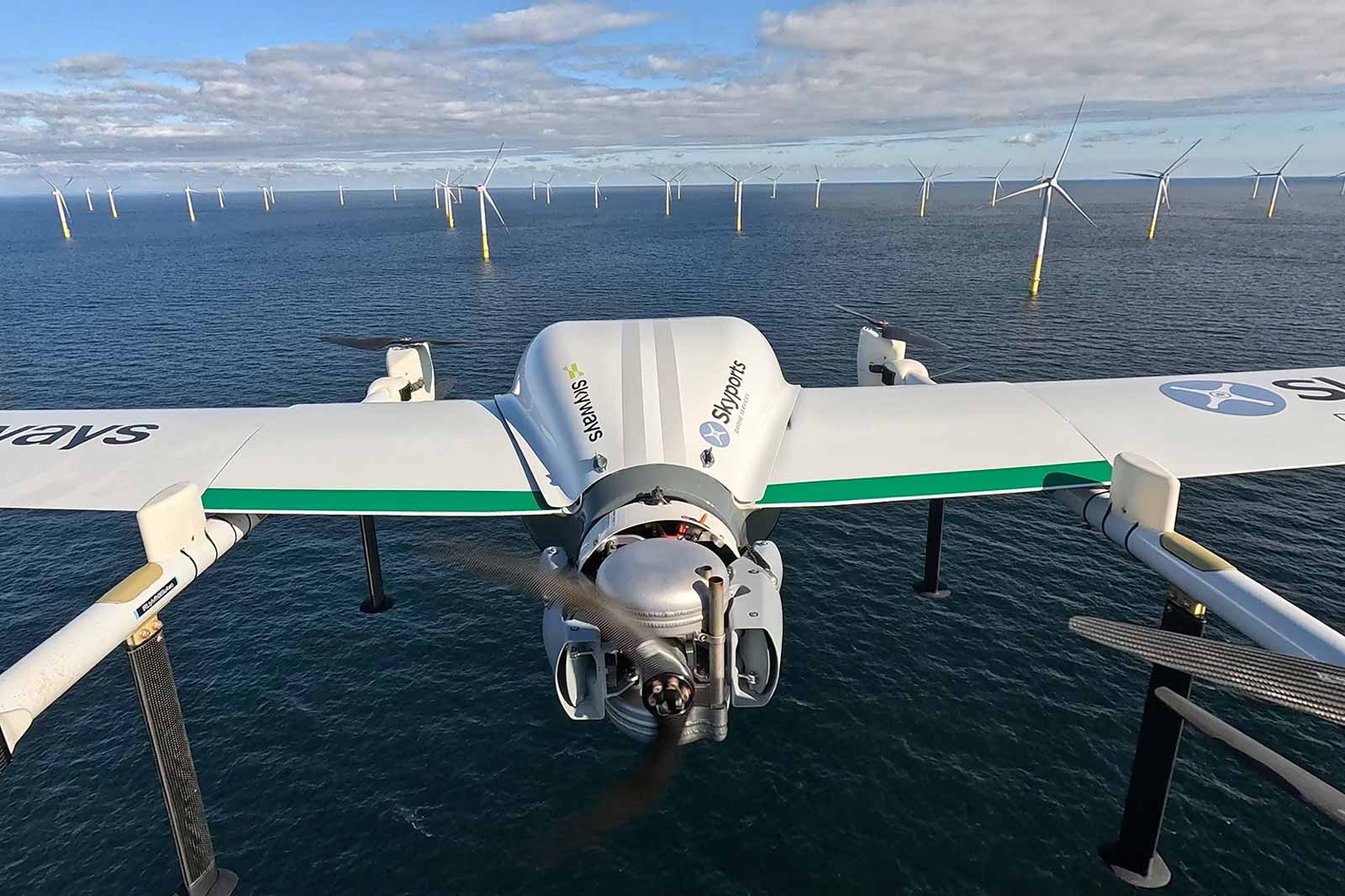 A drone hovers above the water with wind turbines arranged in a grid in the background.