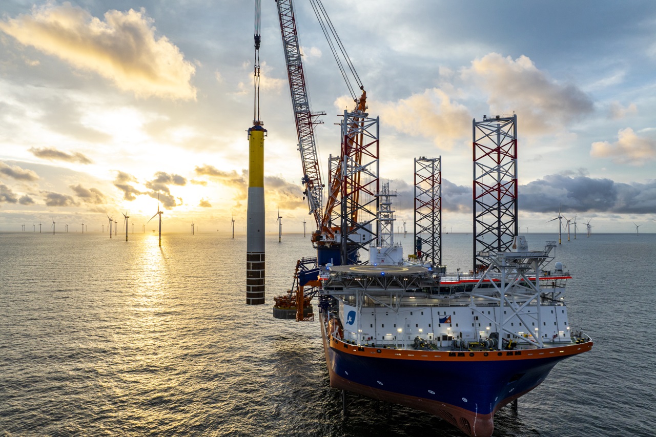 A ship with a crane is lifting a wind turbine at sunset. Additional wind turbines are seen in the distance.
