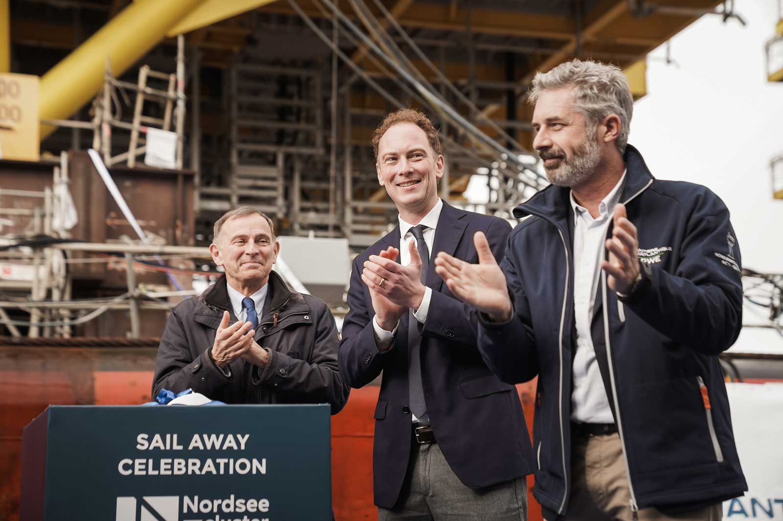 Eine Gruppe von Menschen feiert und klatscht bei einer Abreise-Veranstaltung mit Baumaschinen im Hintergrund.