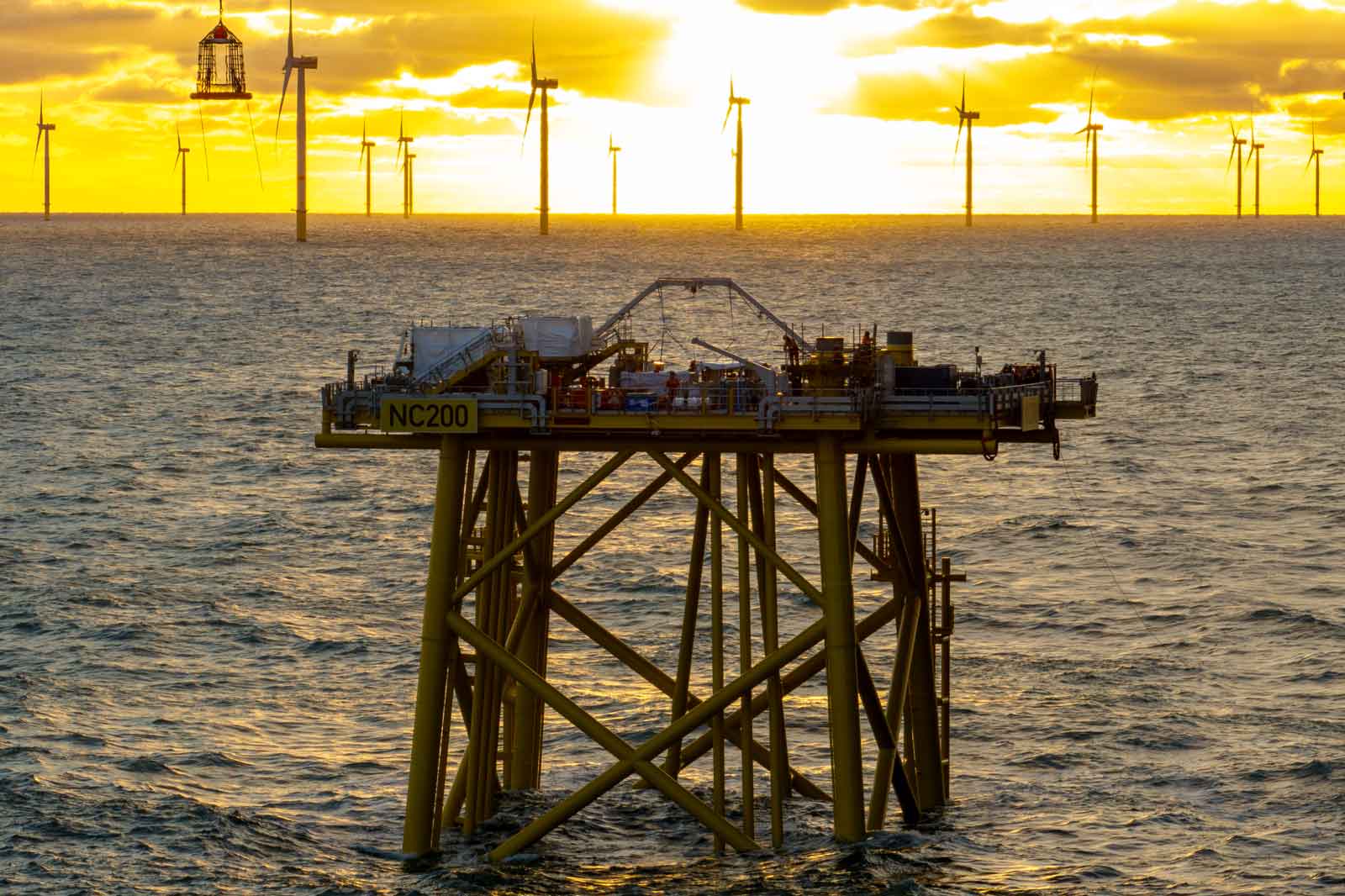 Eine Offshore-Plattform mit der Bezeichnung NC200 steht vor einem Sonnenuntergang, umgeben von Windkraftanlagen.