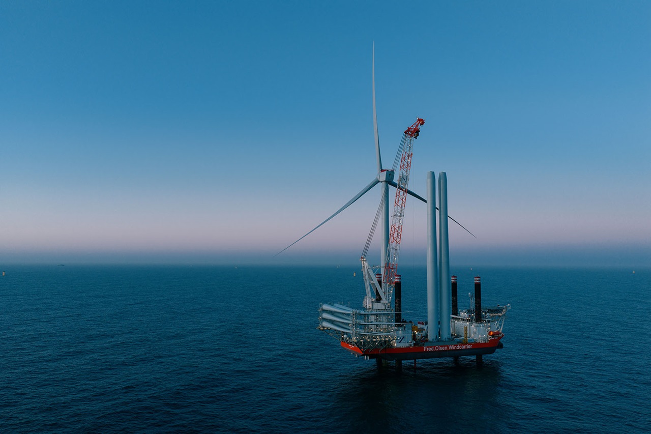 An offshore installation vessel with a crane lifts a wind turbine blade beside assembled turbine towers on a calm sea at dusk.