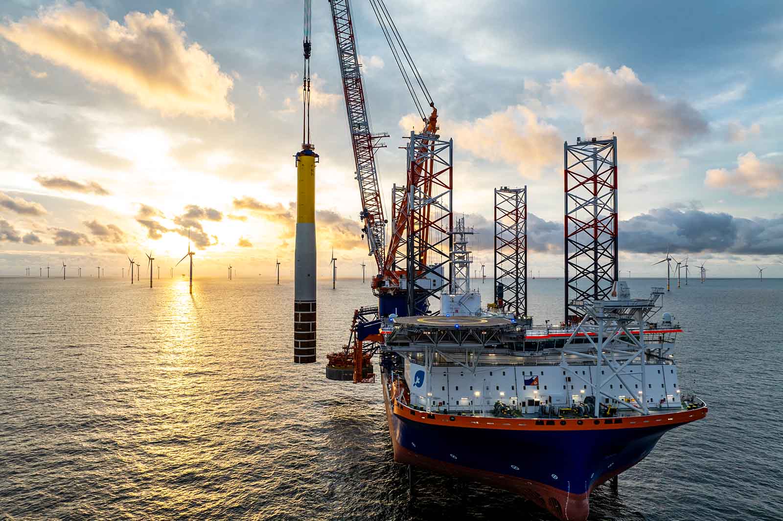 An offshore crane lifts a large wind turbine tower at sunset, beside a large workship with four legs in the sea.