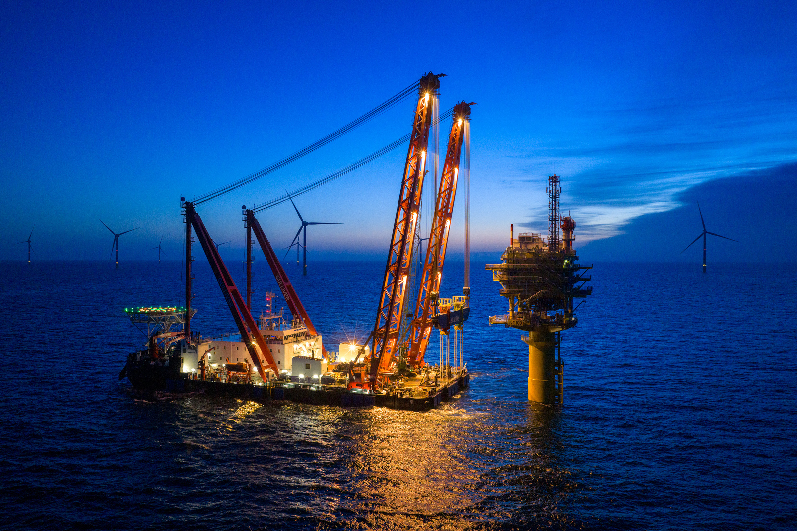 Schwerlastschiff mit Kränen neben beleuchteter Plattform und Windrädern.