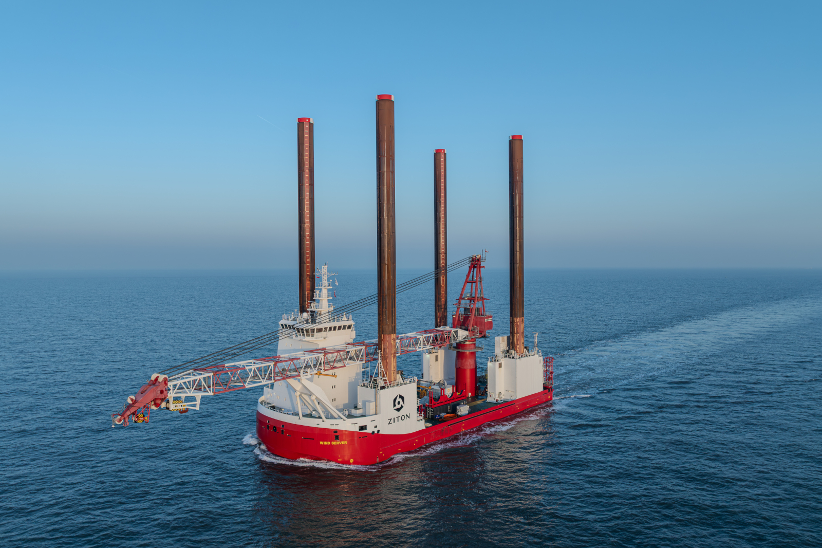 A large red-and-white jack-up vessel with four tall vertical legs and a deck crane moves through calm dark-blue sea under a clear sky.