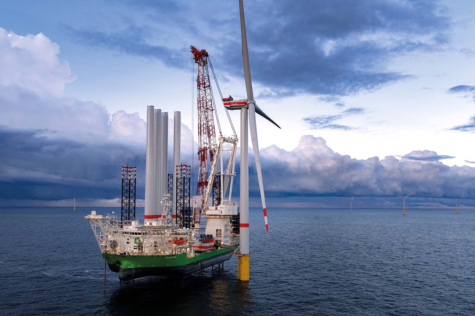 Ein grün gestrichener Versorgungskutter mit weißen Türmen und rot-weiß lackiertem Kran liegt neben einer weißen Offshore-Windturbine über graublauem Meer unter schweren Wolken.