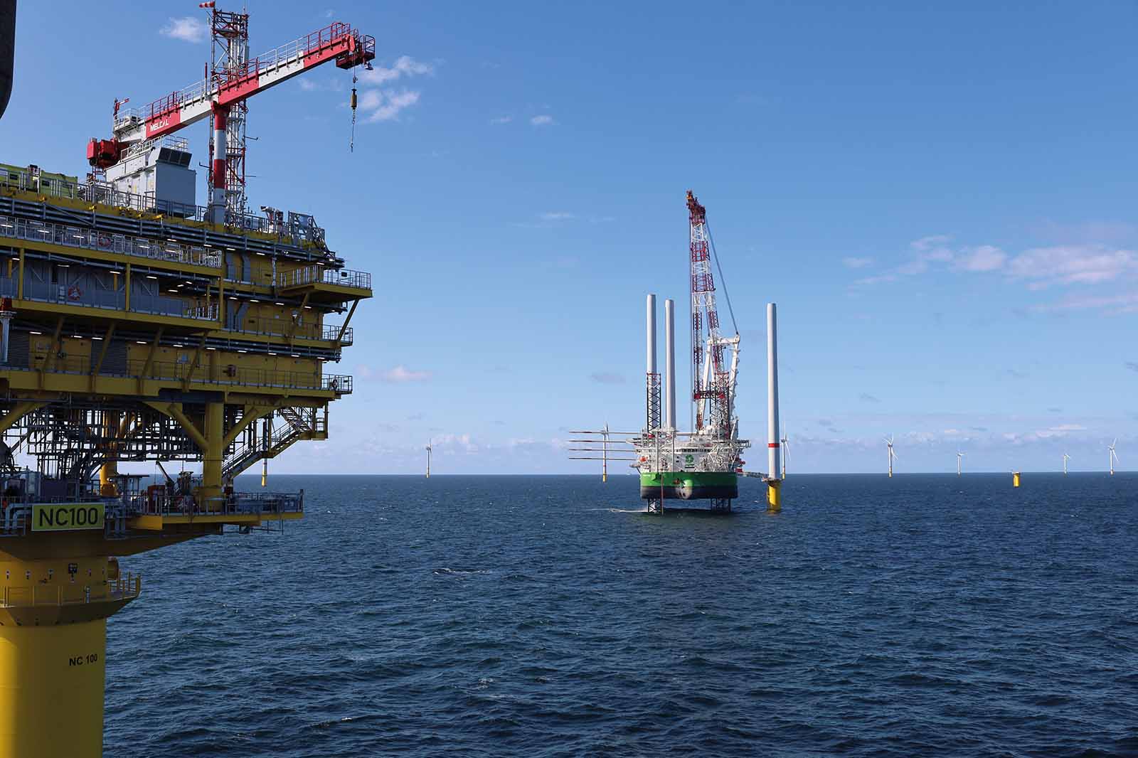 Eine gelbe Plattformstütze mit der Beschriftung „NC100“ dominiert den linken Vordergrund, dahinter steht eine Jack-up-Plattform mit roten Kränen auf ruhigem blauem Meer.