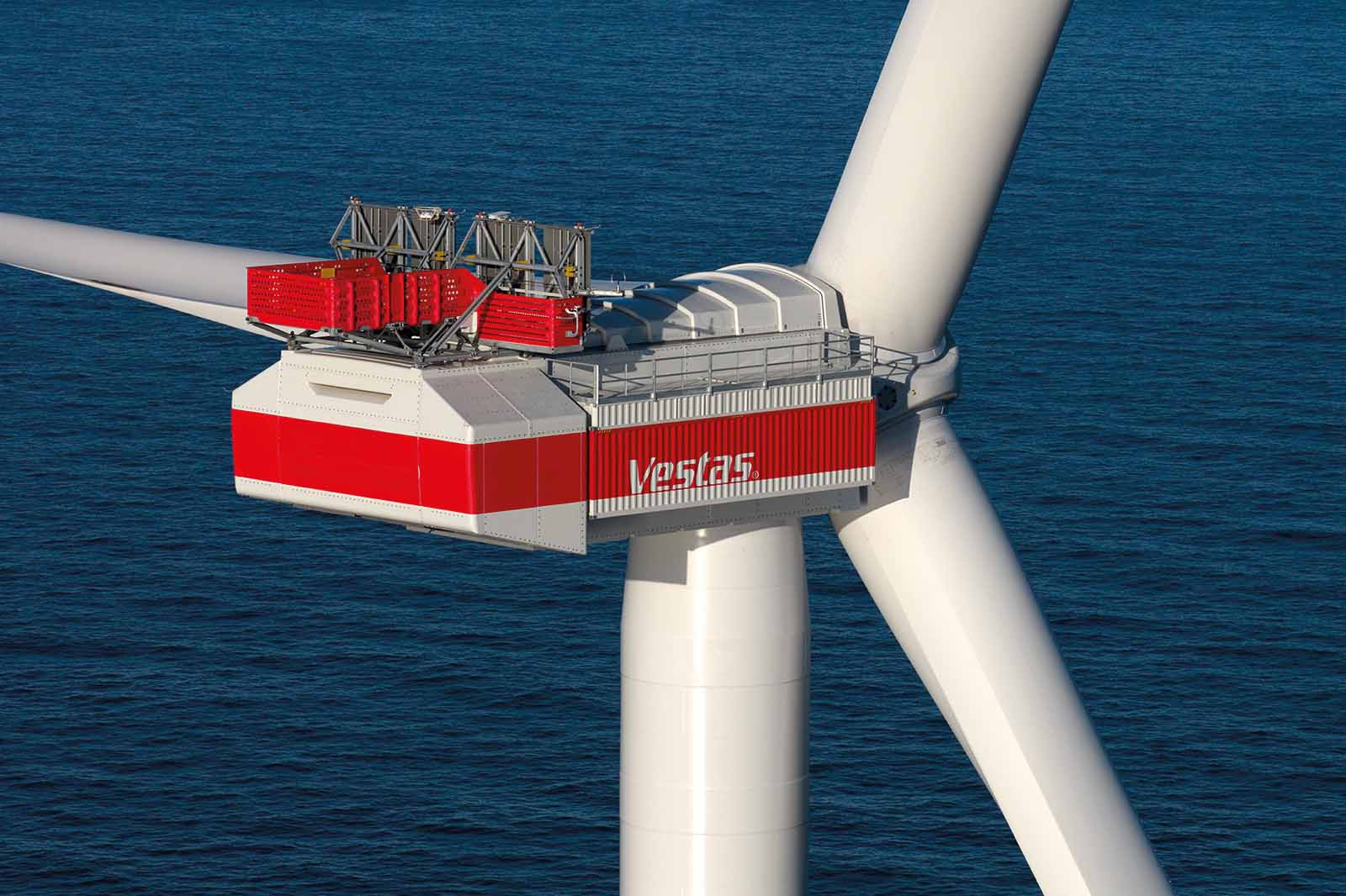 Nahaufnahme einer weißen Windturbinen-Gondel mit roten Verkleidungen und dem Schriftzug „Vestas“ an der Seite, drei Rotorblätter über tiefblauem Meer.