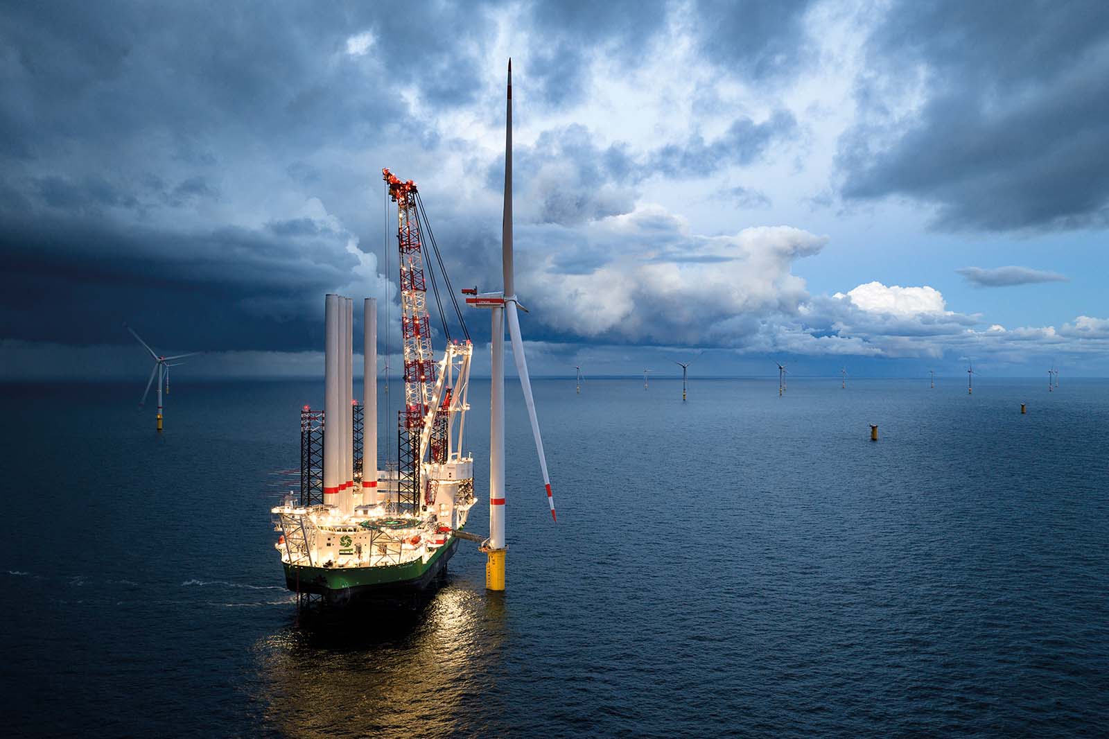 Ein hell beleuchtetes Schiff mit rot-weißen Kränen neben einer weißen Windturbine, die aus dunklem blauem Meer unter stürmischen Wolken ragt.