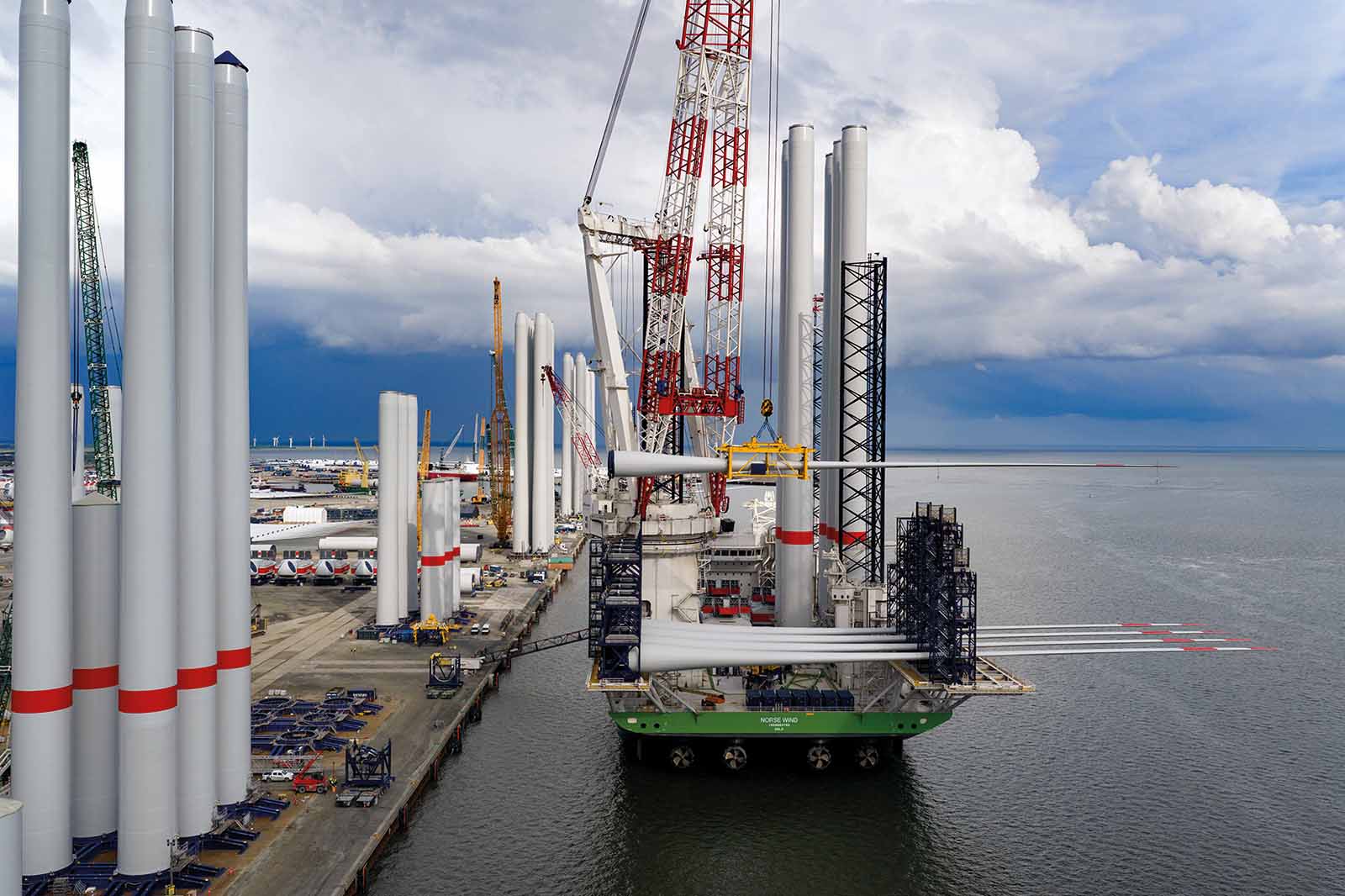 Ein grünes Schwerlastschiff am Betonkai transportiert weiße zylindrische Turmsegmente unter roten Kränen vor einem dramatischen wolkenverhangenen Himmel.