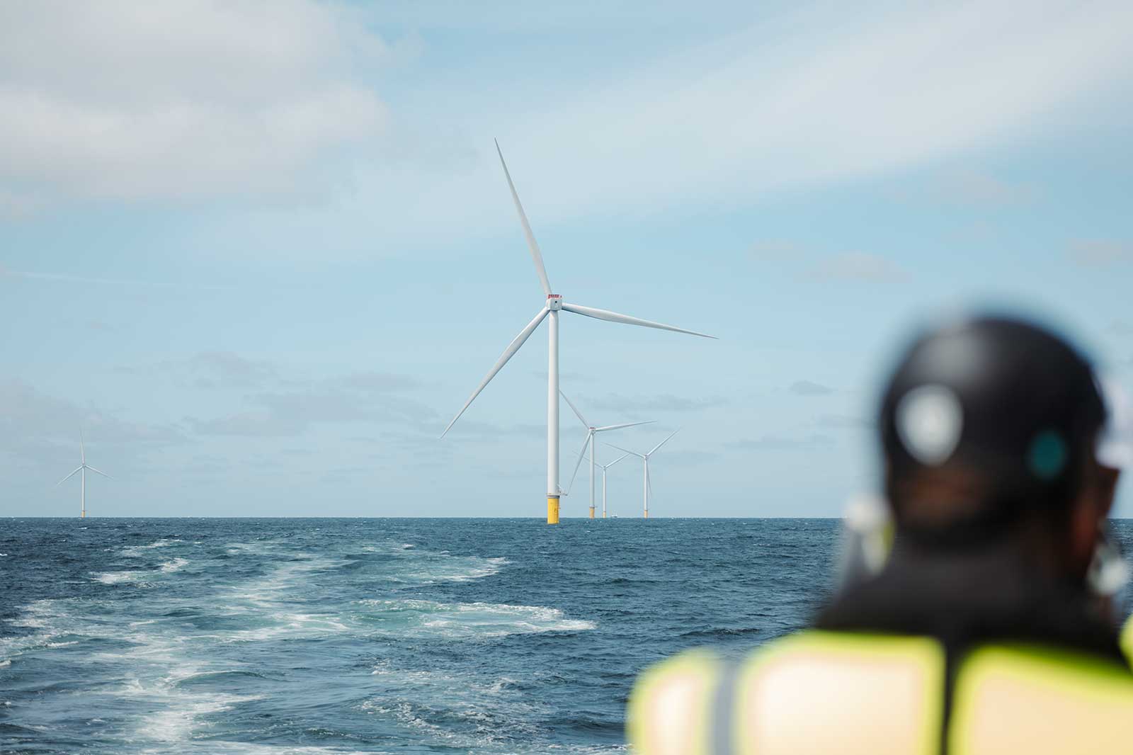 A figure in a fluorescent life jacket and black helmet occupies the right foreground, looking toward white offshore wind turbines above a deep blue sea.