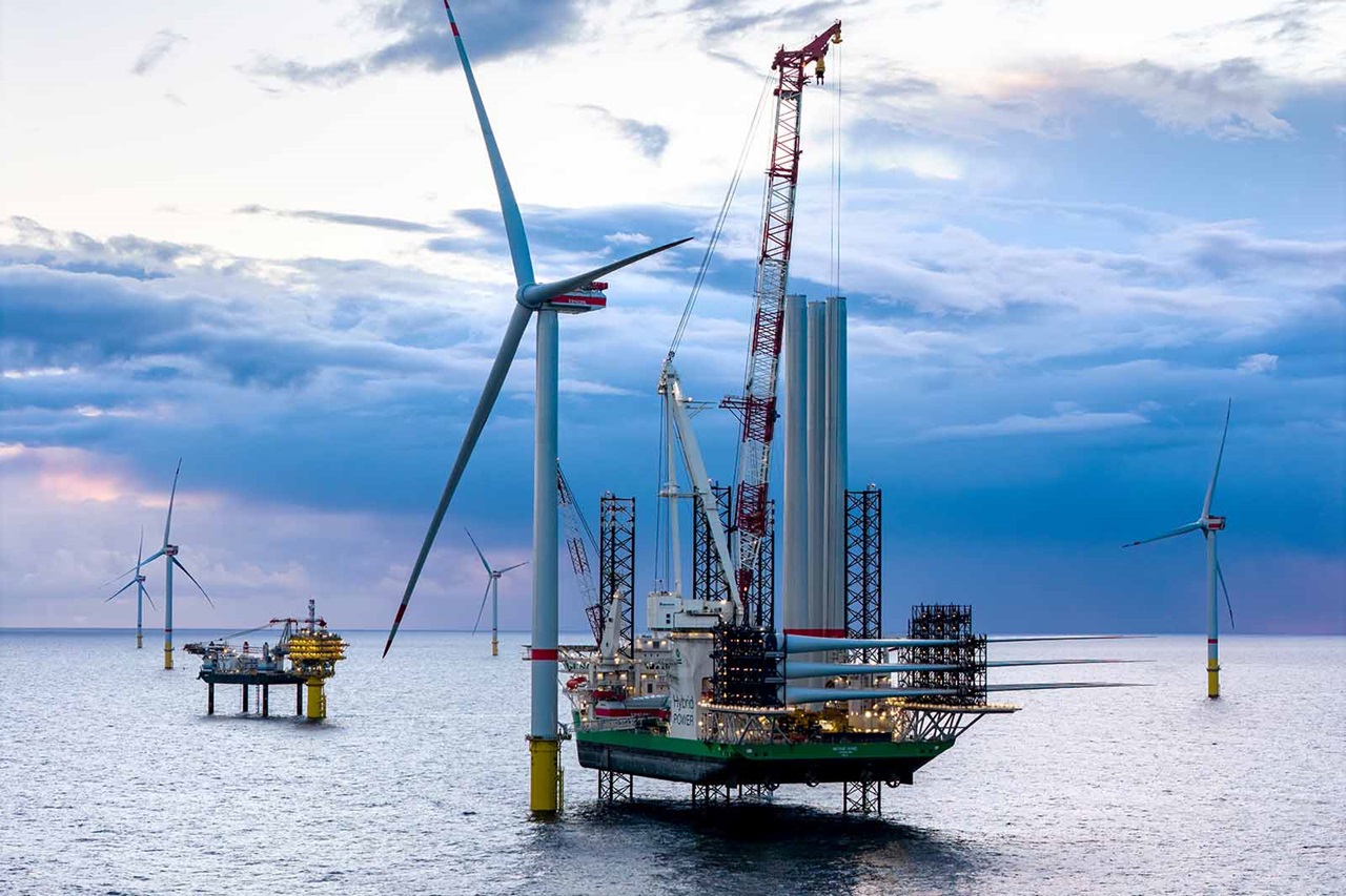 Ein kranbestücktes Service-Schiff mit Gerüst und einer Arbeitsplattform steht zwischen weißen Offshore-Windturbinen in einem ruhigen grau-blauen Meer unter bewölktem Himmel.