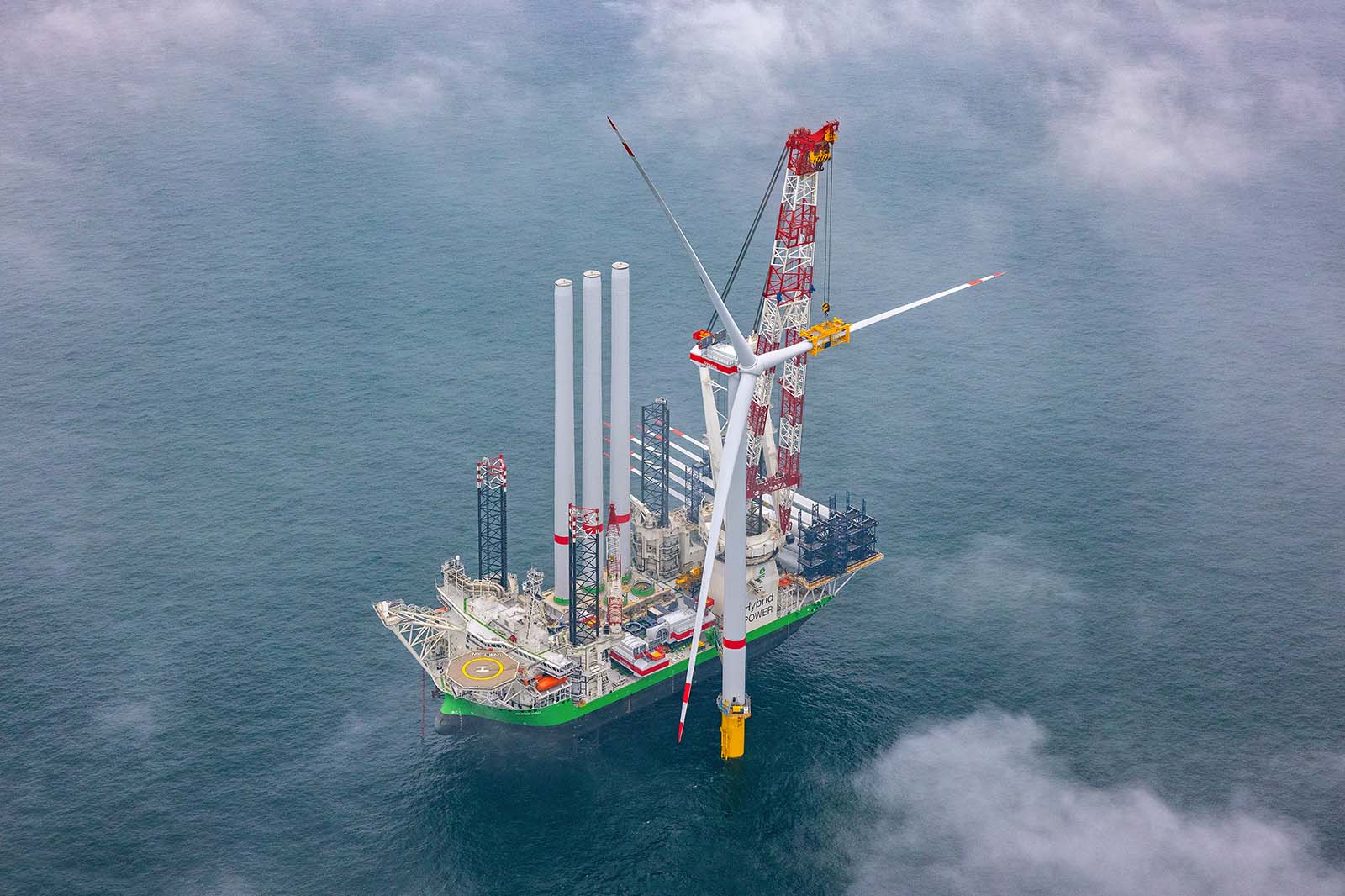 Ein Kran auf einem grün gestrichenen Installationsschiff hebt einen weißen Windturbinenmast mit dreiflügeligem Rotor über dem ruhigen grauen Meer. Rote Kranstreben und aufgestapelte stählerne Rohrsegmente liegen auf dem Deck unter einem nebligen Wolkenhimmel.
