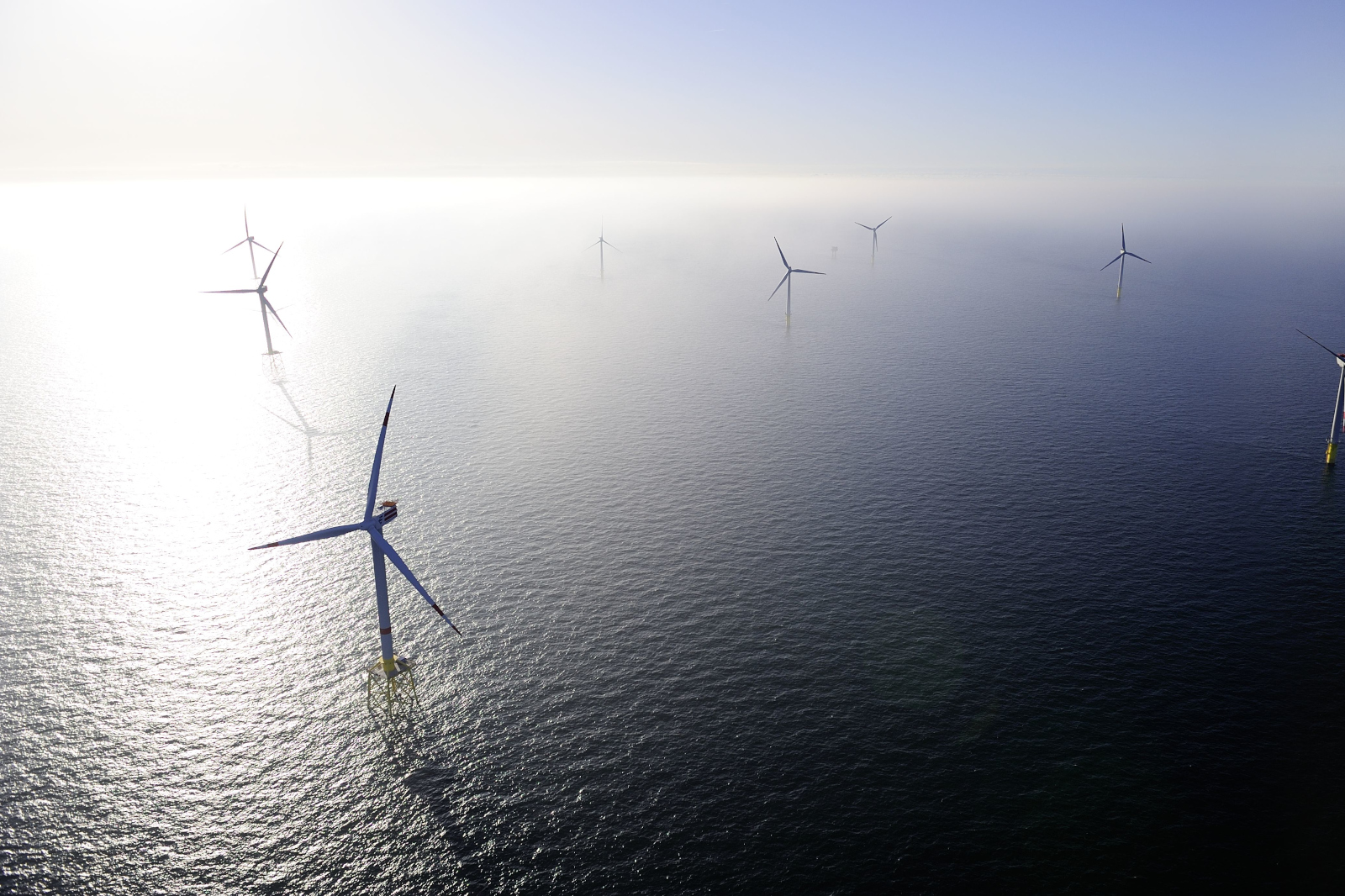 Mehrere weiße Offshore-Windkraftanlagen stehen über ruhiger, blaugrauer See im Morgennebel, ihre hohen Türme und langen Rotorblätter werfen schlanke Schatten auf schimmerndes Wasser.