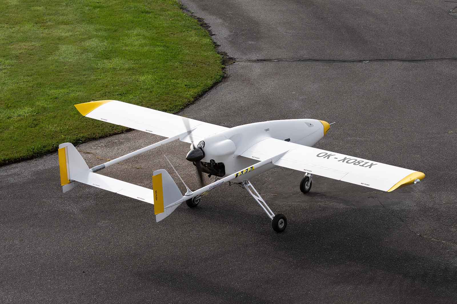 Ein weißes Leichtflugzeug mit Doppelleitwerk, gelben Flügelspitzen und der Kennung „OK-1005X” ist ruhig auf dunklem Rollfeld neben grünem Gras geparkt.