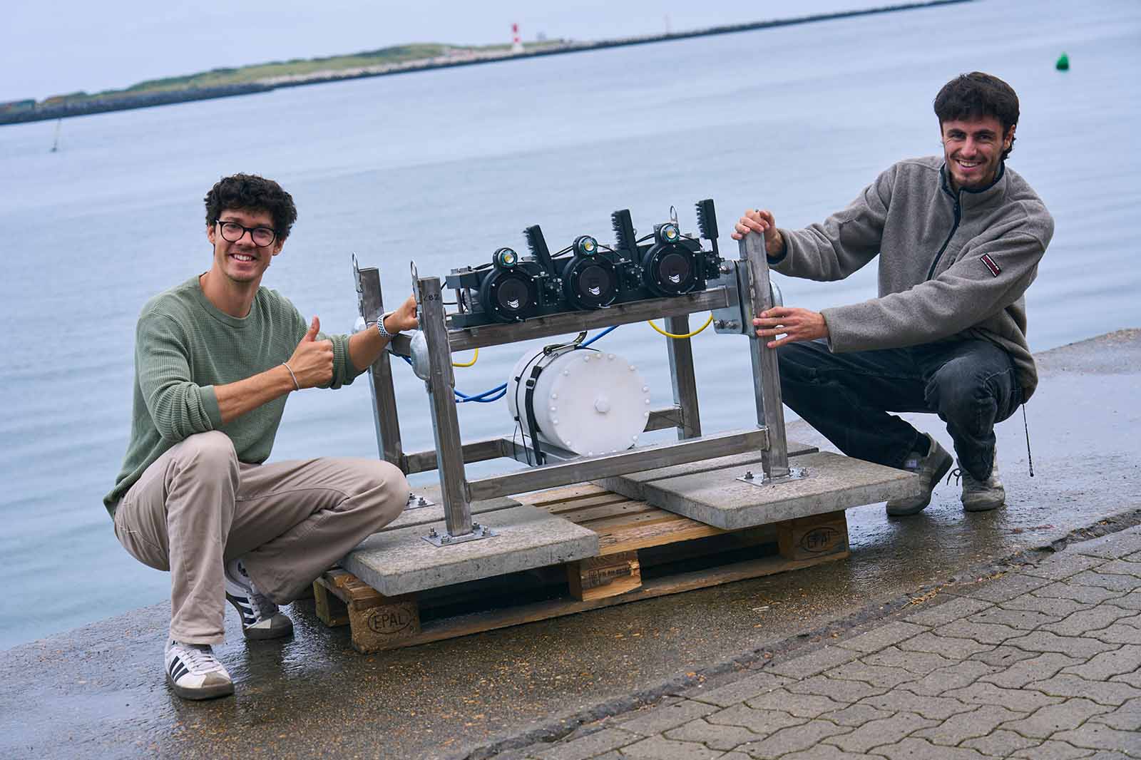 Zwei junge Männer knien am Hafen vor graublauem Wasser neben einem silbrig-metallischen Gestell auf einer Holzpalette und lächeln.