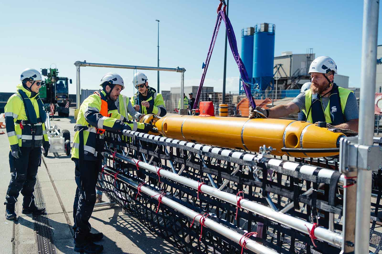 Drei Personen in weißen Schutzhelmen und gelben Warnjacken arbeiten konzentriert an einem orangefarbenen Metallzylinder auf einem schwarzen Gestell am Betonkai vor blauen Silos.