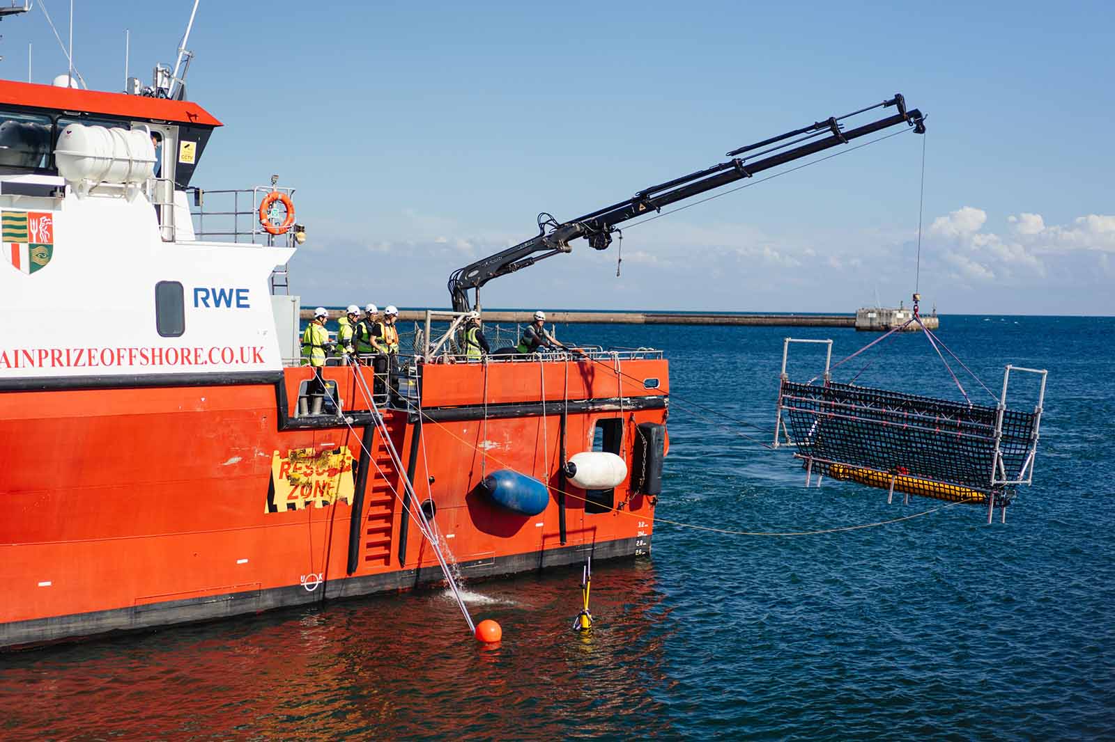 Orangefarbenes Arbeitsschiff hebt Metallgitterkorb mit schwarzem Kran über blaues Meer. Besatzung in gelben Warnwesten steht auf dem Deck, und der Schriftzug „RWE“ ist sichtbar.