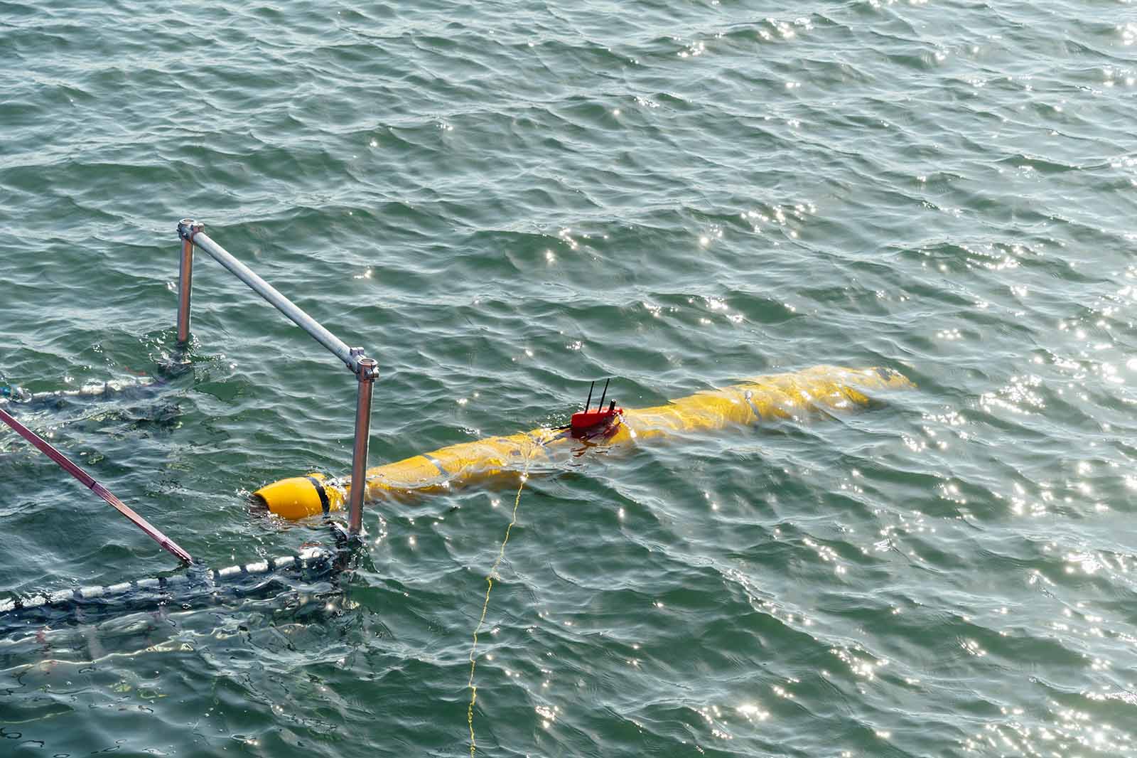 Gelber torpedoförmiger Messkörper schwimmt halb unter grünlichem, welligem Wasser, an dem ein rotes Elektronikmodul mit Antennen, ein Metallgestänge und ein Seil befestigt sind.