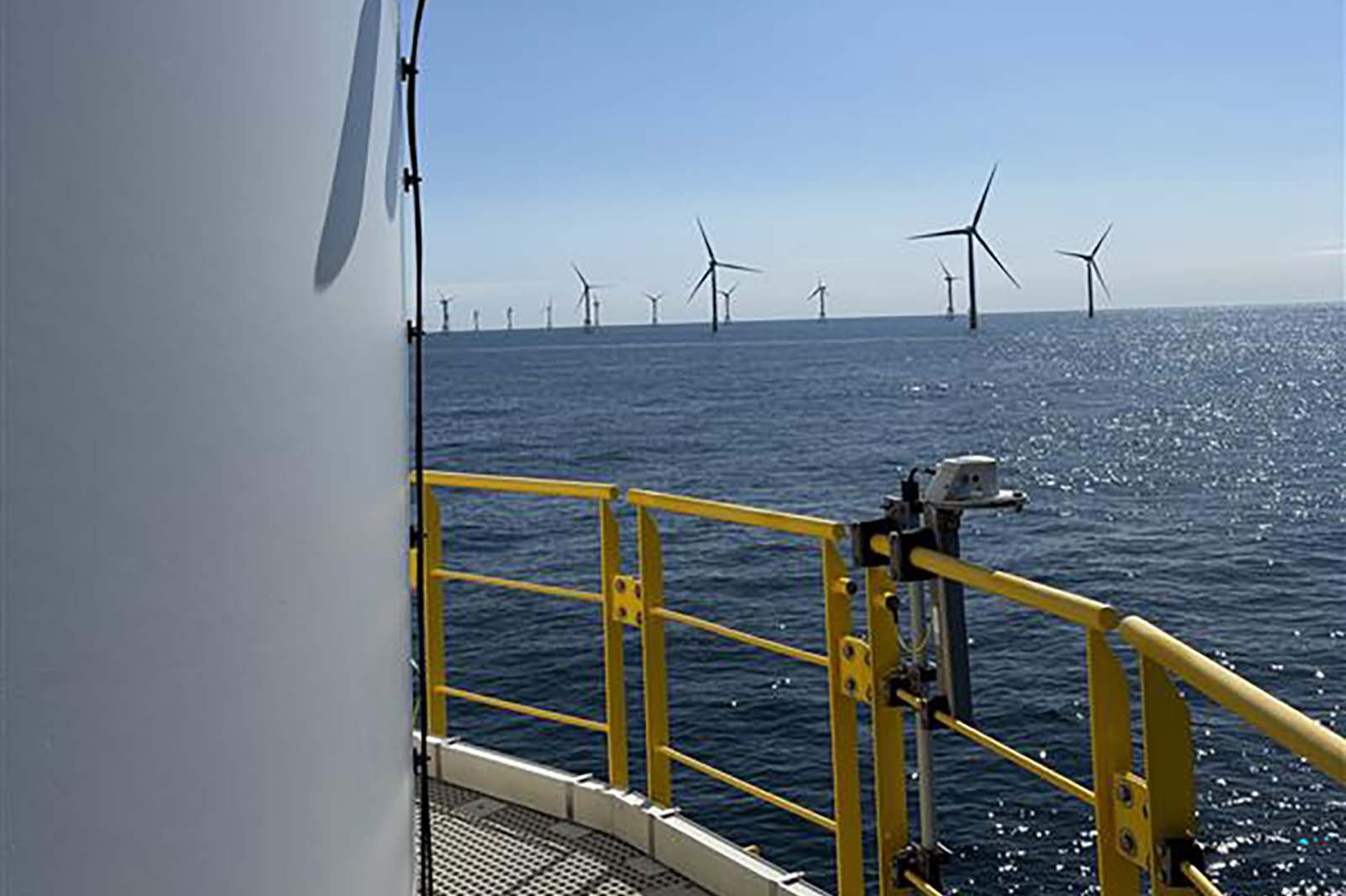 Ein gelbes Metallgeländer und ein metallisches Gitterdeck säumen die weiße Turmwand einer Offshore-Windkraftanlage, dahinter breitet sich blaues Meer mit Windrädern am Horizont aus.