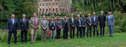 Eine Gruppe von Menschen in formeller Kleidung, die im Freien stehen, mit grüner Umgebung und einem historischen Gebäude im Hintergrund.