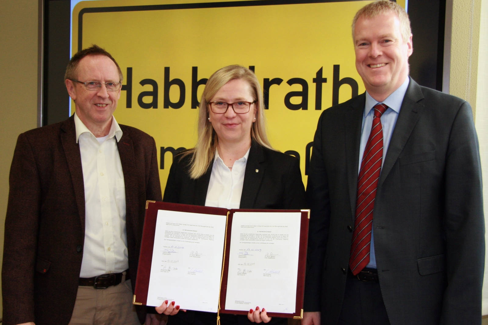 Drei Personen halten Dokumente und stehen vor einem gelben Schild mit Text. Formelle Kleidung und eine professionelle Umgebung.