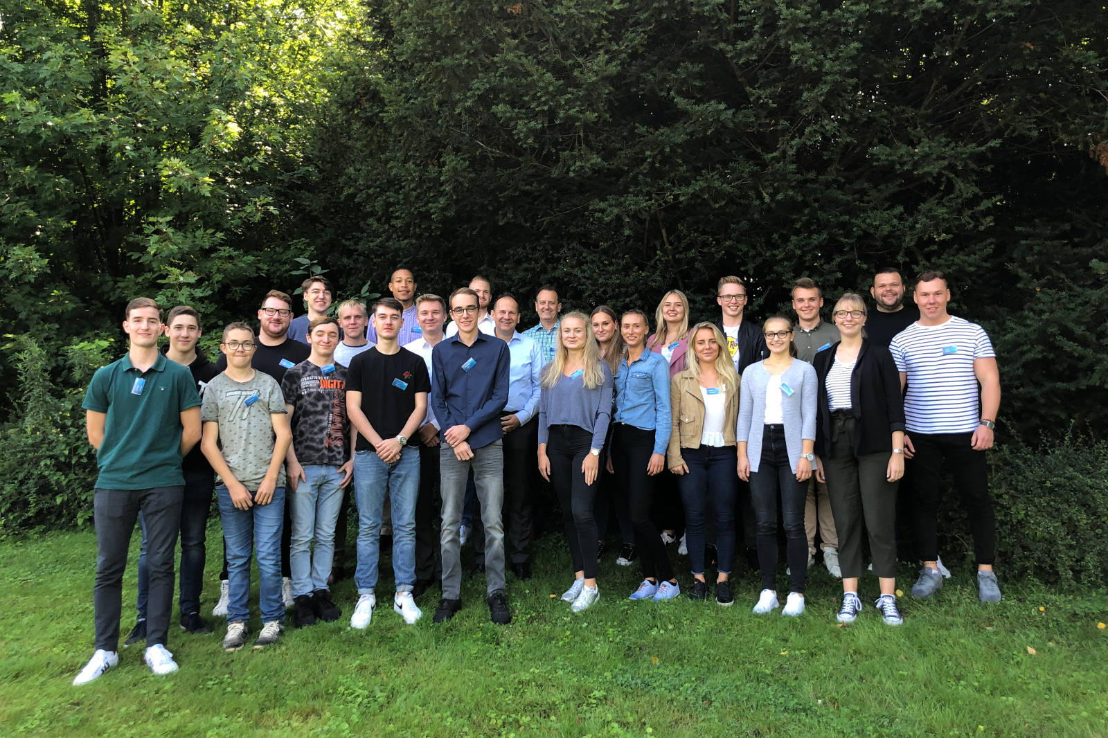 Eine Gruppe von Menschen steht in einem üppigen grünen Bereich, alle tragen Namensschilder, mit Bäumen im Hintergrund.