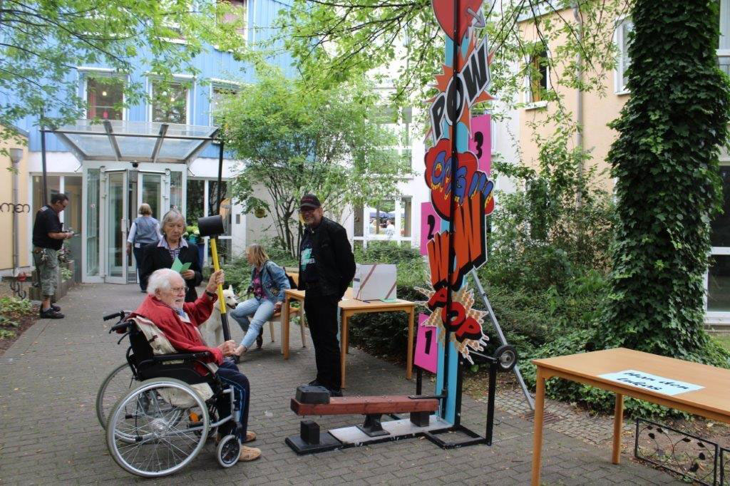 Eine lebendige Außenansicht mit einem Spielaufbau und einem großen bunten Schild. Mehrere Personen sind damit beschäftigt.