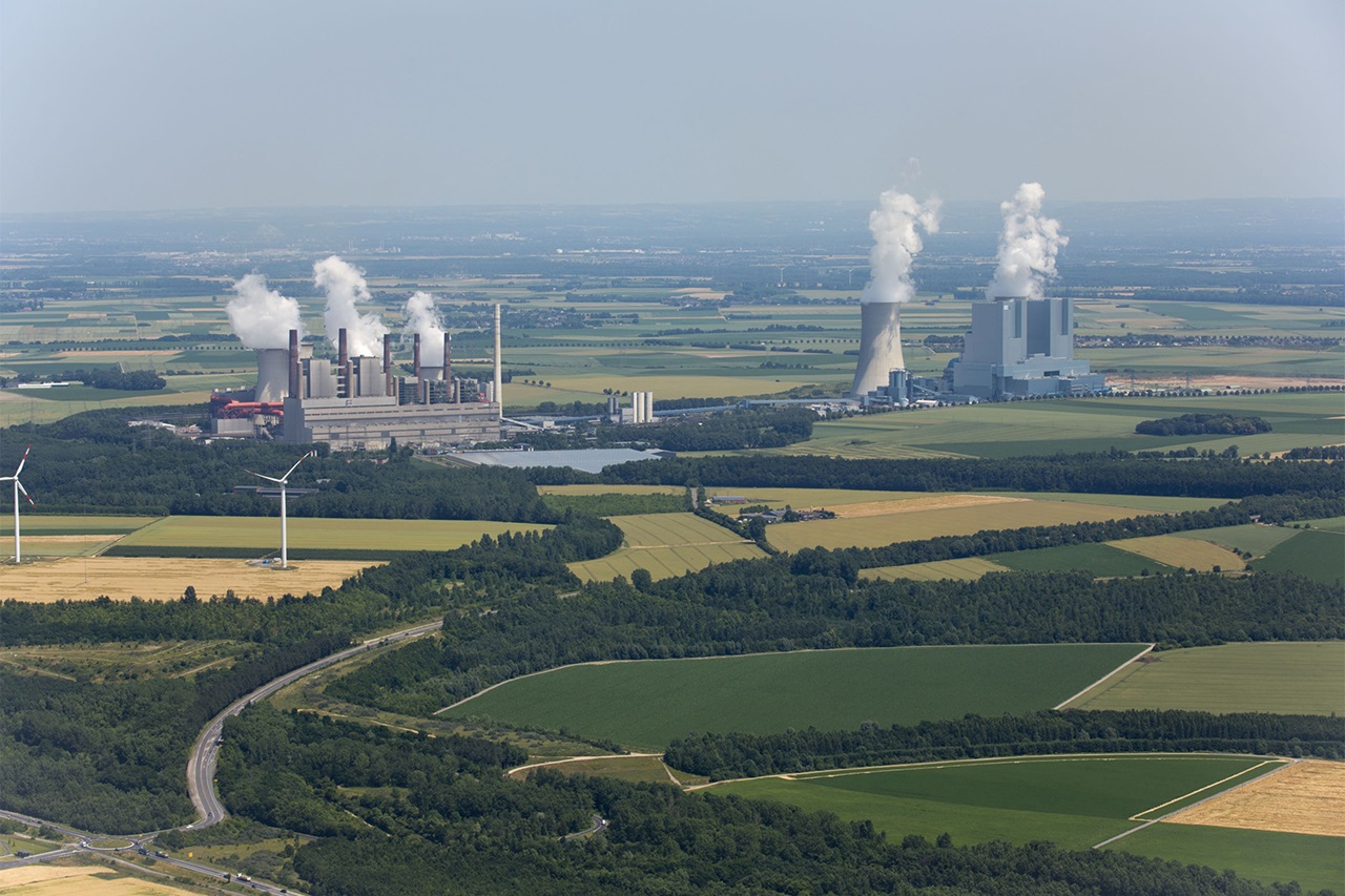 Luftaufnahme von Industrie-Kraftwerken, die Dampf abgeben, umgeben von Feldern und Windkraftanlagen.